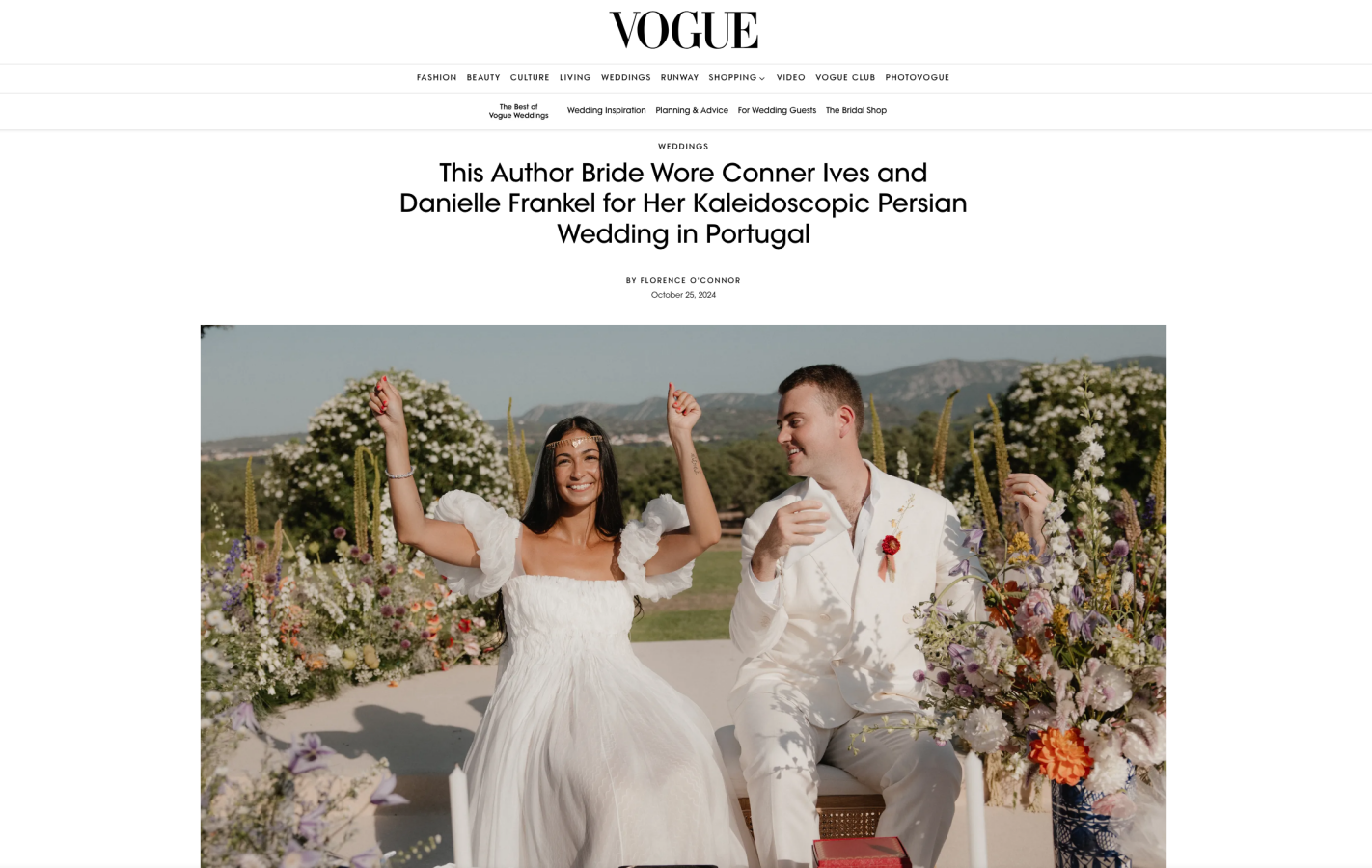 A smiling bride and groom sit outdoors during their wedding ceremony, surrounded by flowers, with scenic mountains in the background.
