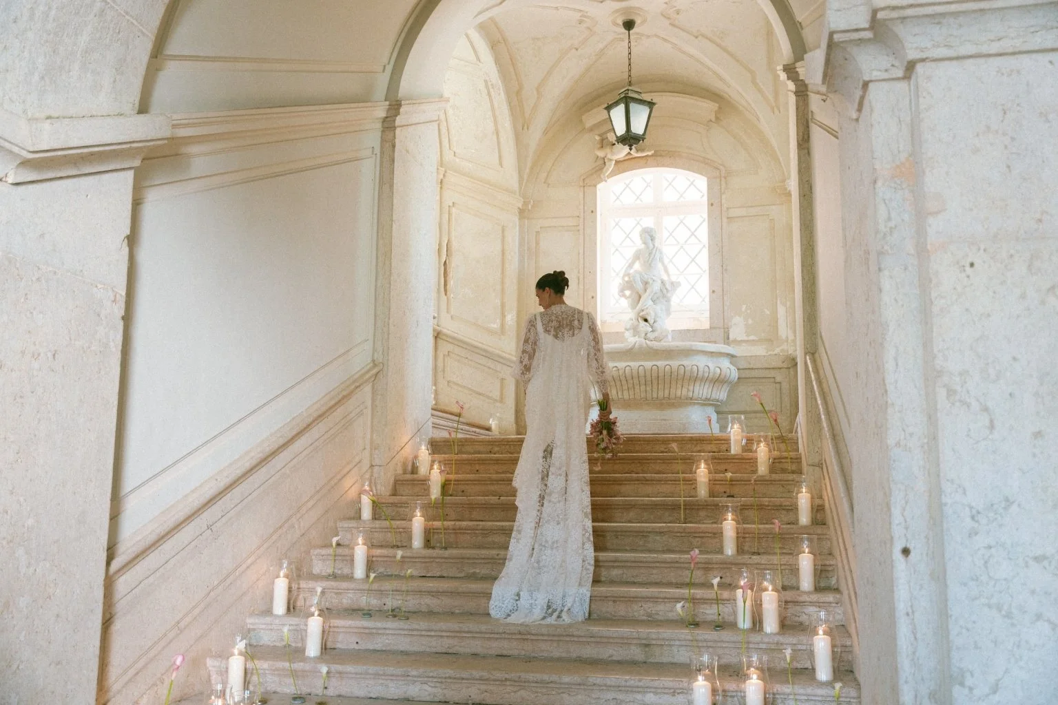A woman in a white lace wedding dress stands on marble stairs inside a historic building, surrounded by candles and flowers, with a statue and large window behind her.