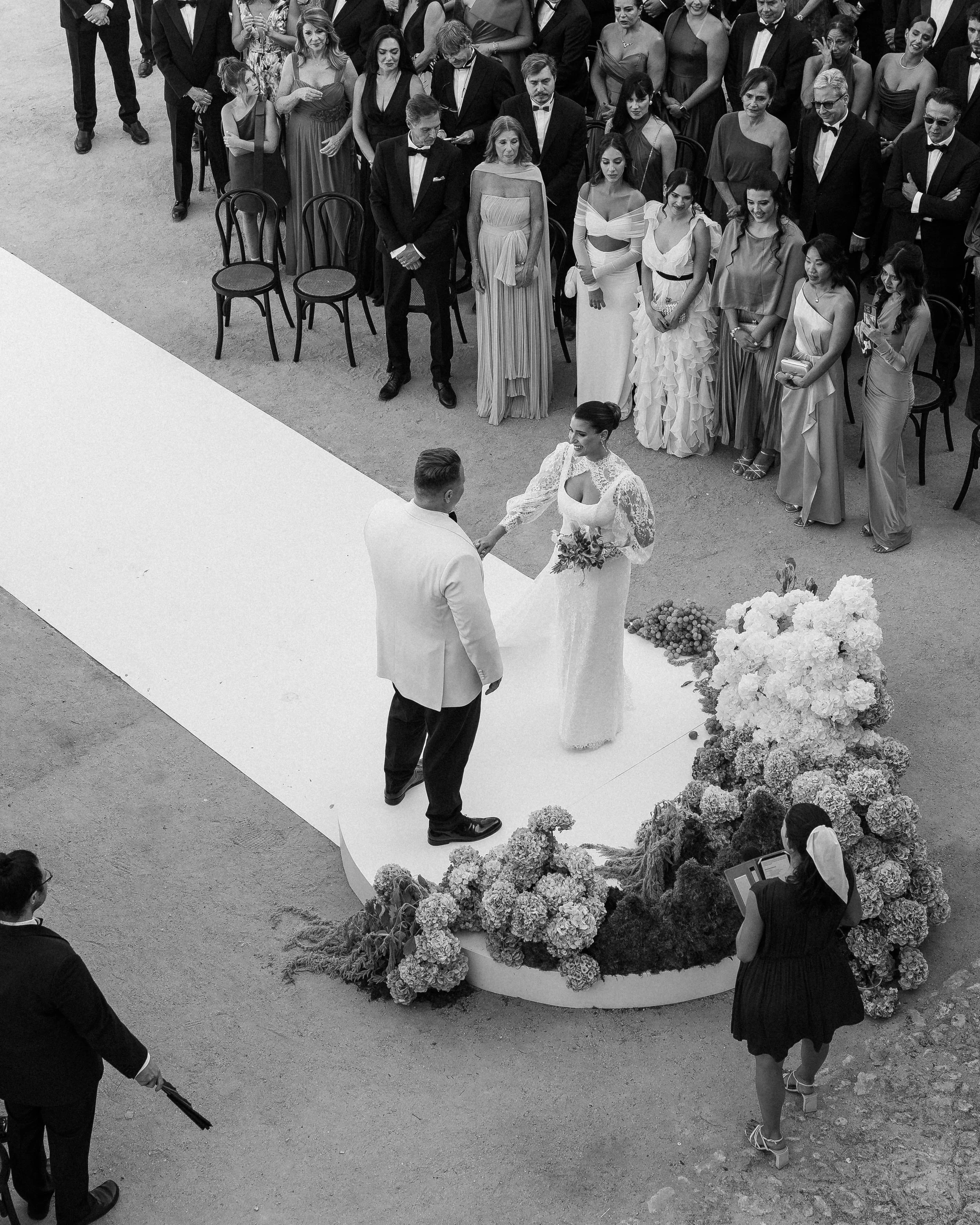 A black and white photo of a wedding ceremony with the bride and groom holding hands in front of a minimalist backdrop, surrounded by floral arrangements, and guests standing behind them in formal attire.