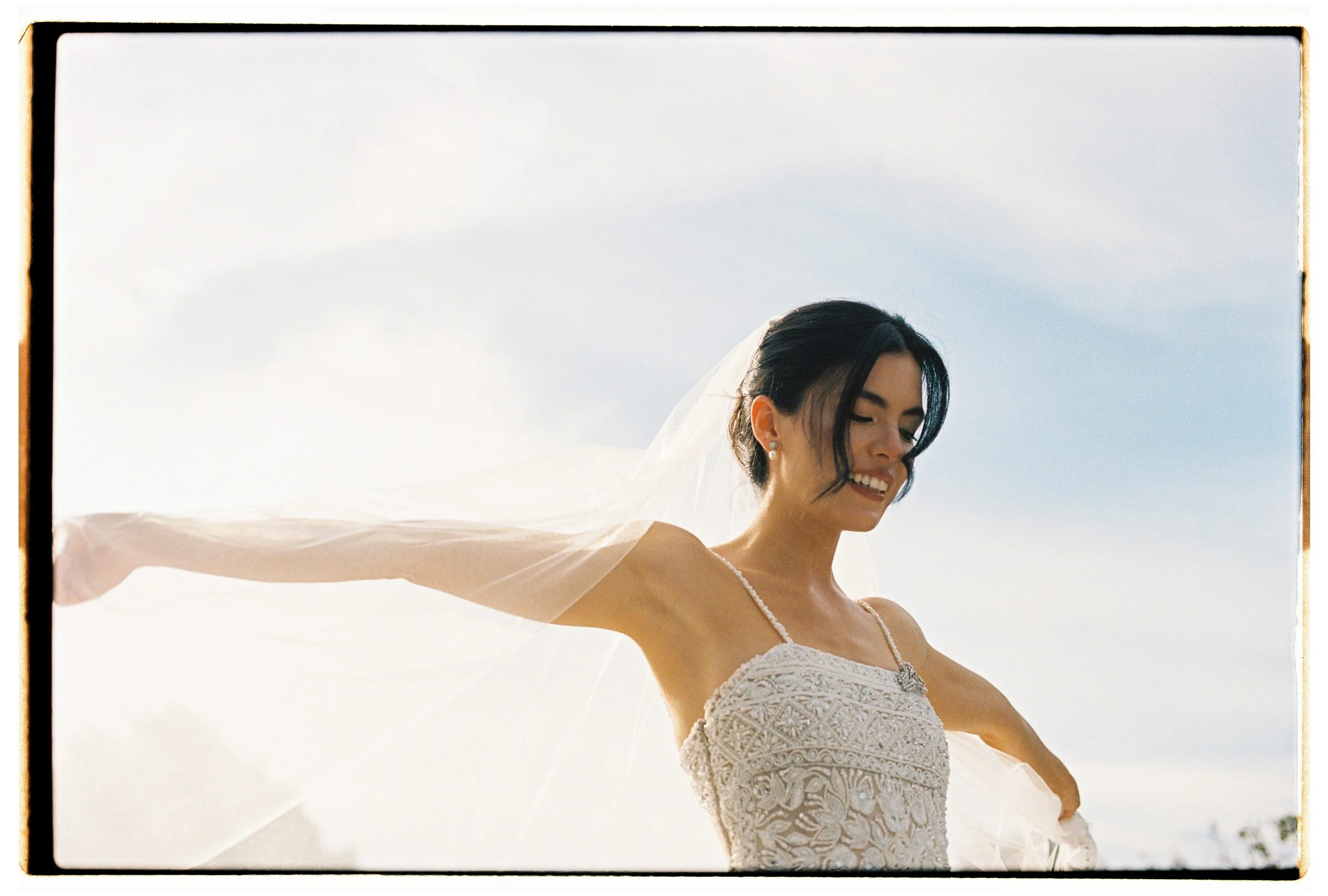 A woman in a wedding dress with a veil smiling with arms outstretched outdoors during daytime.