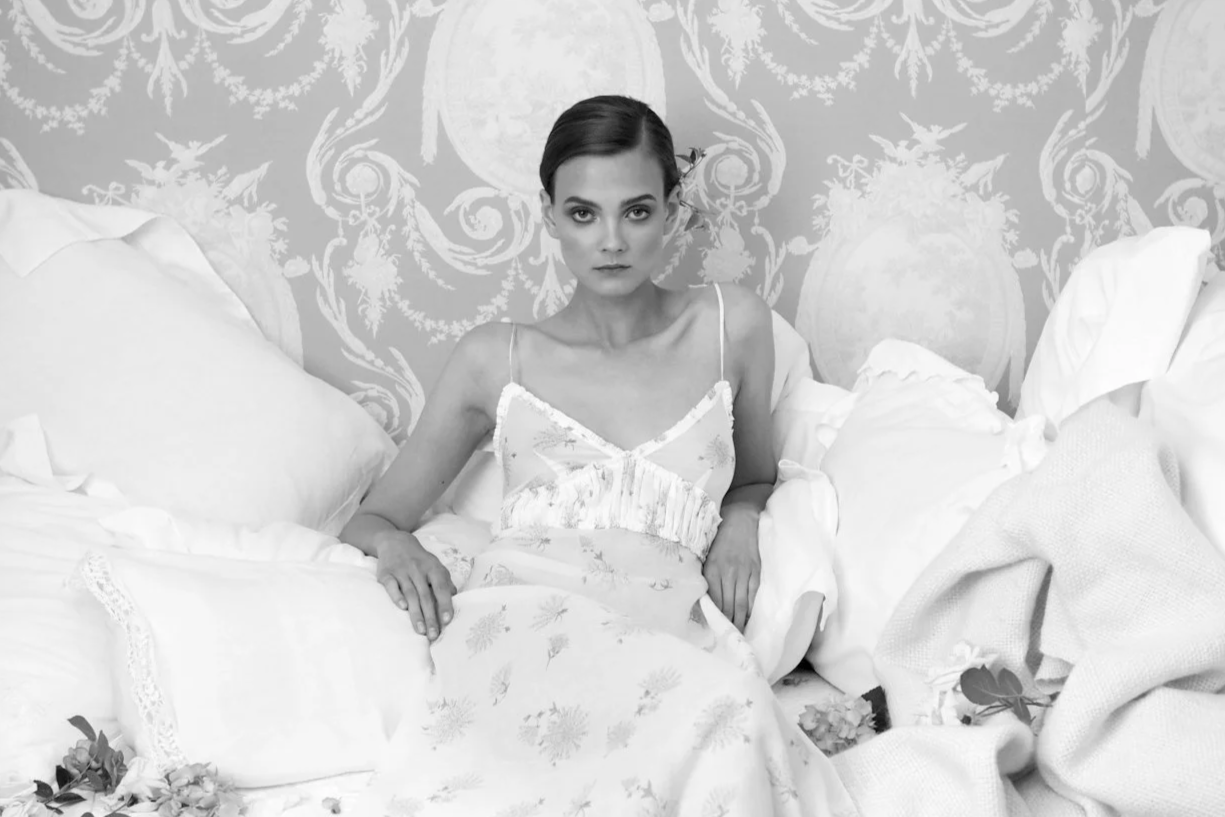 A woman sitting on a bed surrounded by pillows, with a vintage floral wallpaper background. She has short, dark hair and is wearing a light-colored, sleeveless dress.