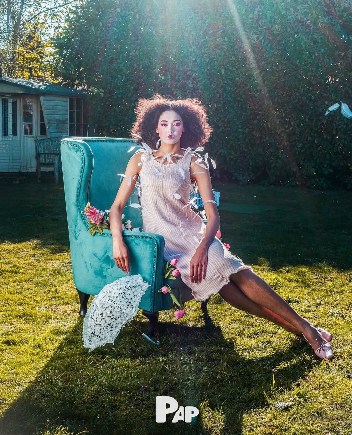 Fashion editorial styling: Model with dark curls seated on a vintage turquoise armchair amidst wildflowers and sunlight.