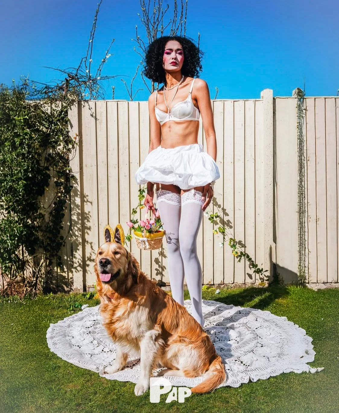 Whimsical garden fashion: Model in white lingerie and stockings with a dog in bunny accessories, styling assistance by Mara Bernemann.