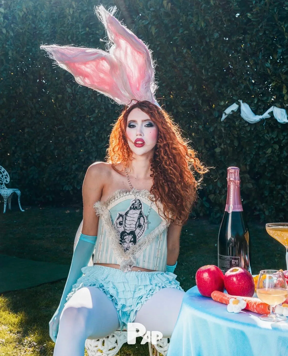 Conceptual styling: Model with red curls and bunny ears in a surreal outdoor dining scene with blue gloves and lacy accents.
