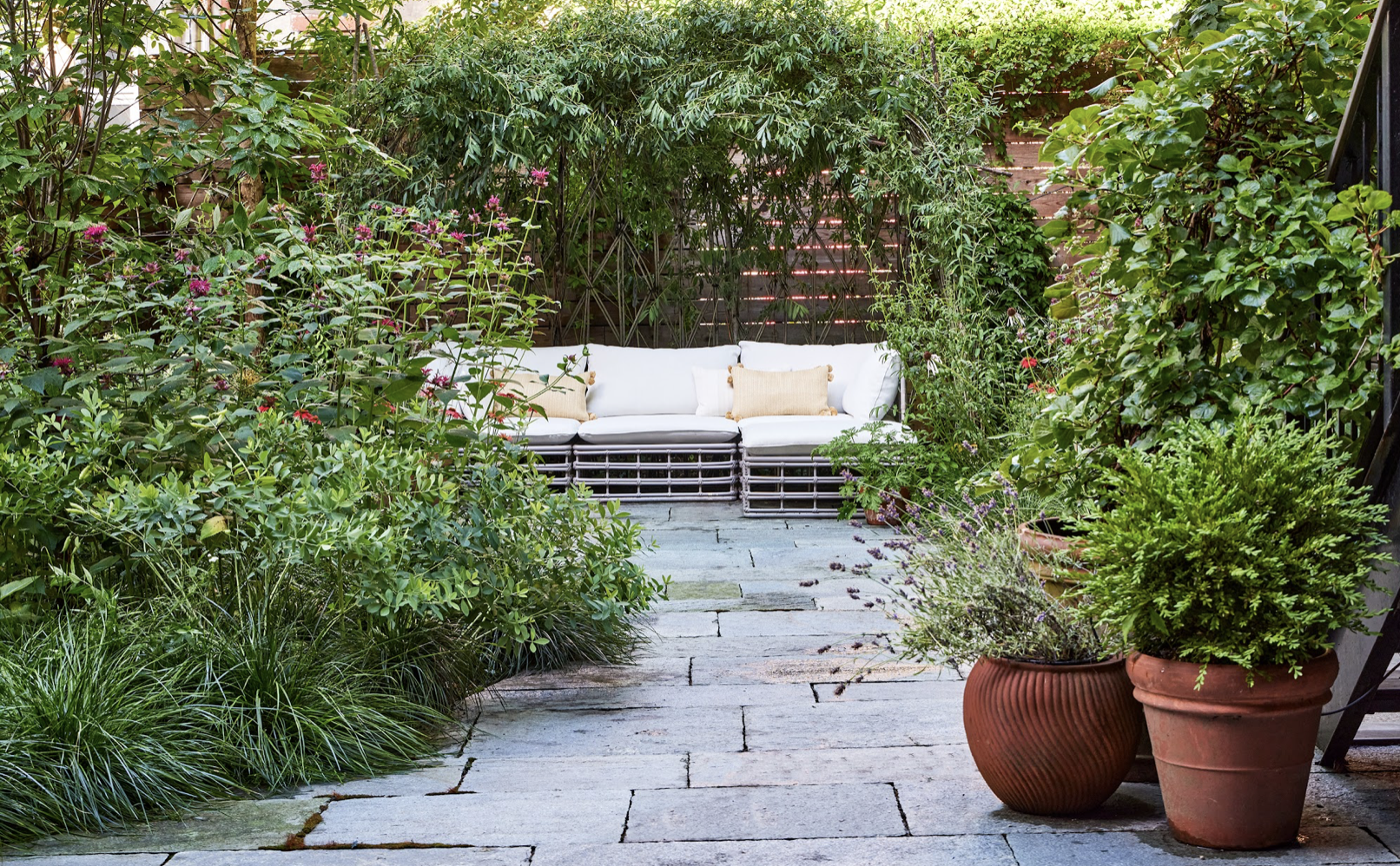 A serene backyard patio with stone paving, lush green plants, flowering bushes, and a white outdoor sofa with cream pillows in the background.