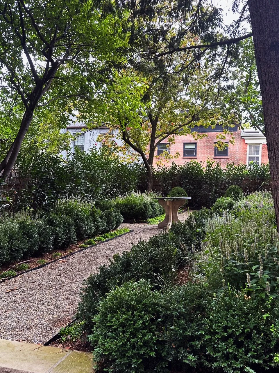 A lush garden with a gravel pathway, green bushes, and a birdbath, surrounded by trees and a brick house in the background.