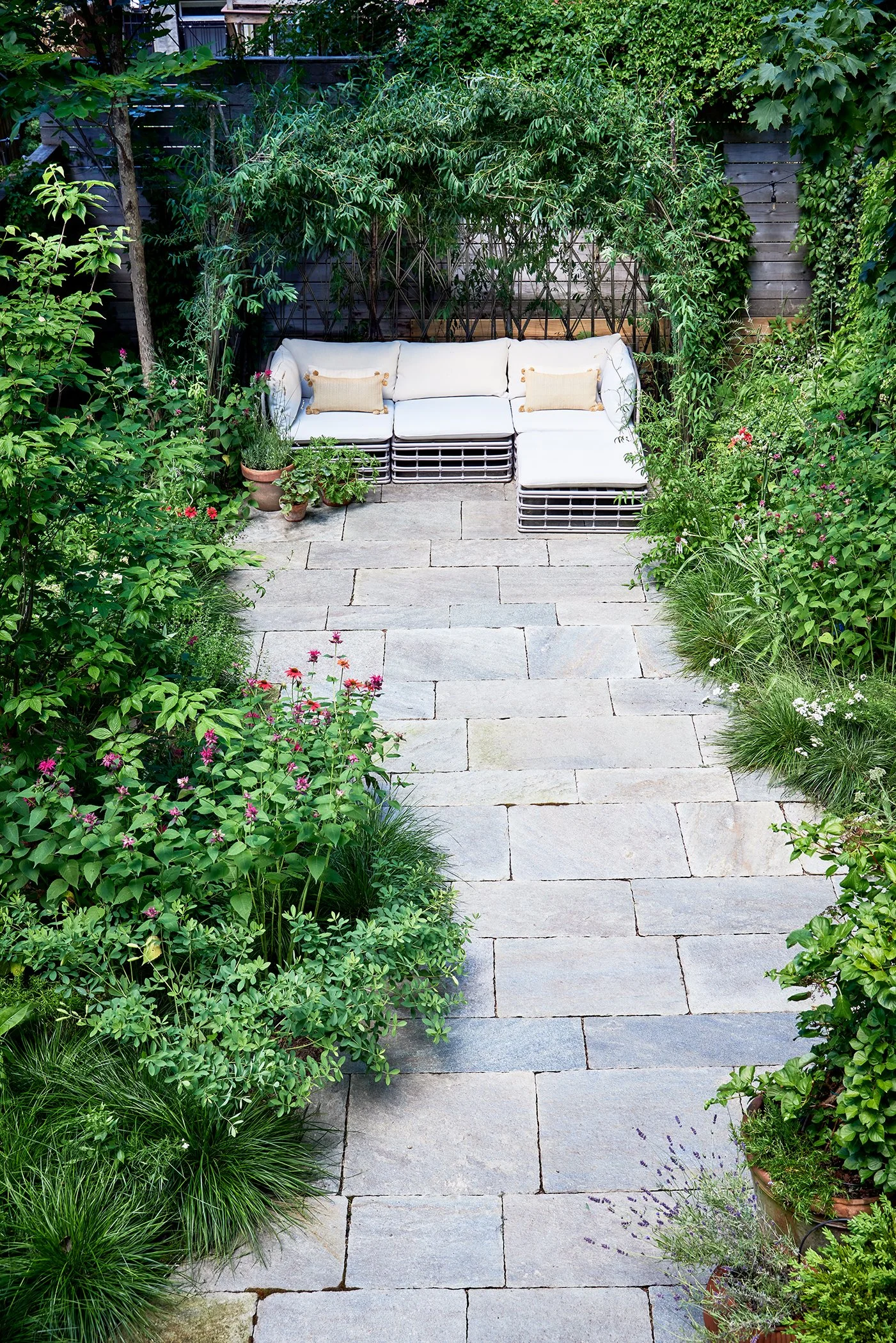 A garden patio with a stone pathway leading to a white outdoor sofa with two beige pillows, surrounded by lush green plants and flowers, and a wooden fence in the background.