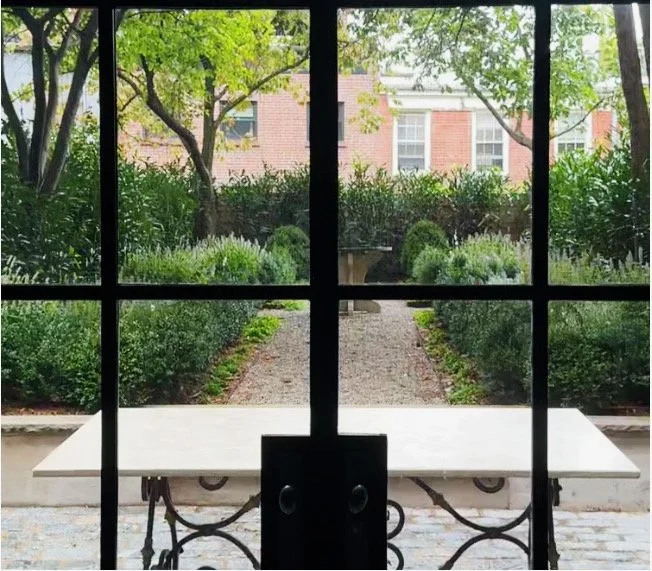 View through a glass window showing a garden with a path, bushes, and trees, with a brick building in the background.