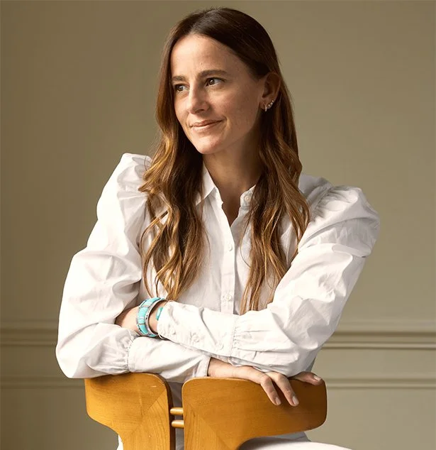 A woman with long, wavy brown hair wearing a white blouse, sitting on a wooden chair with a neutral background.