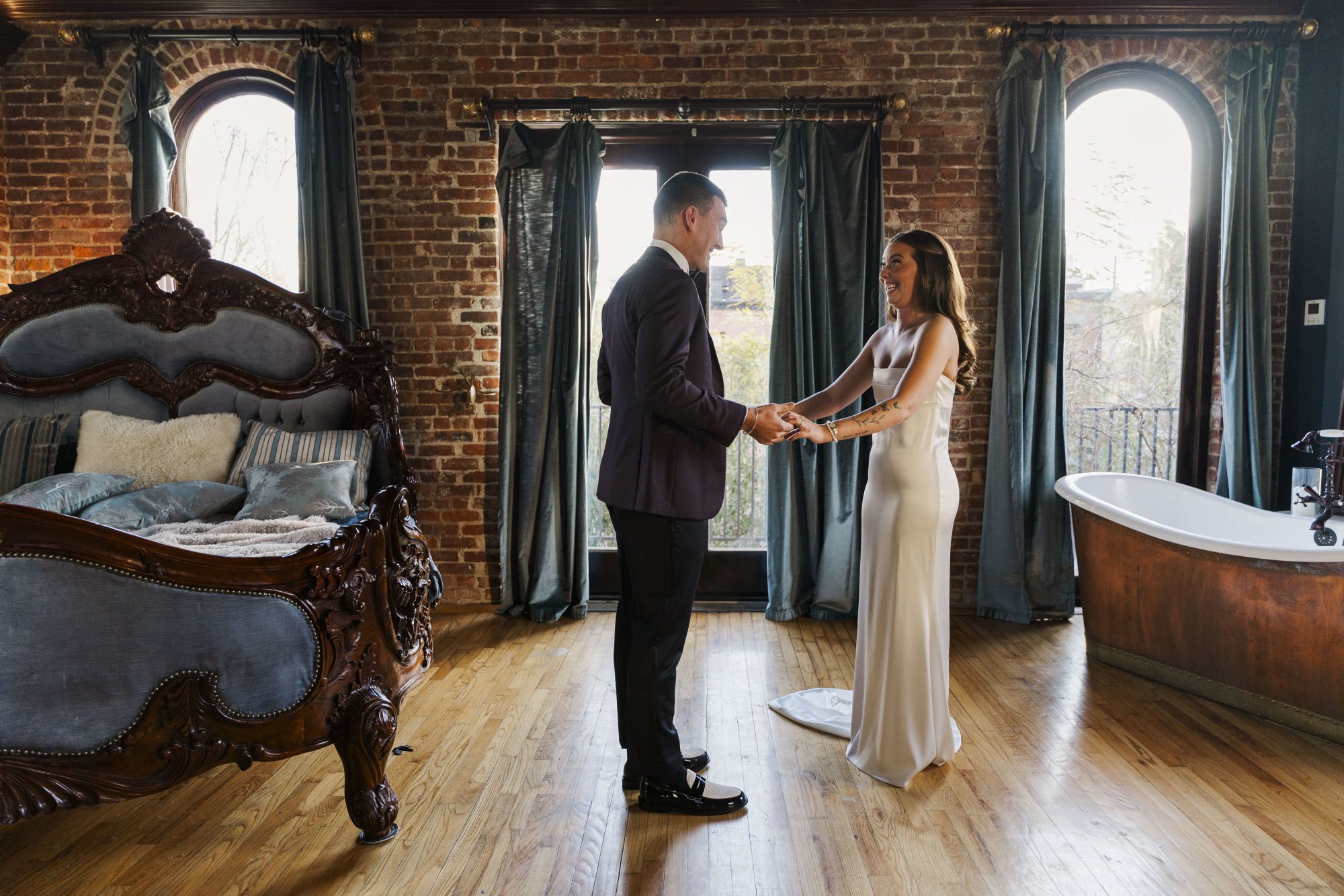 A man and woman holding hands and smiling at each other in a rustic bedroom, with vintage furniture, brick walls, large windows with curtains, and a clawfoot bathtub.