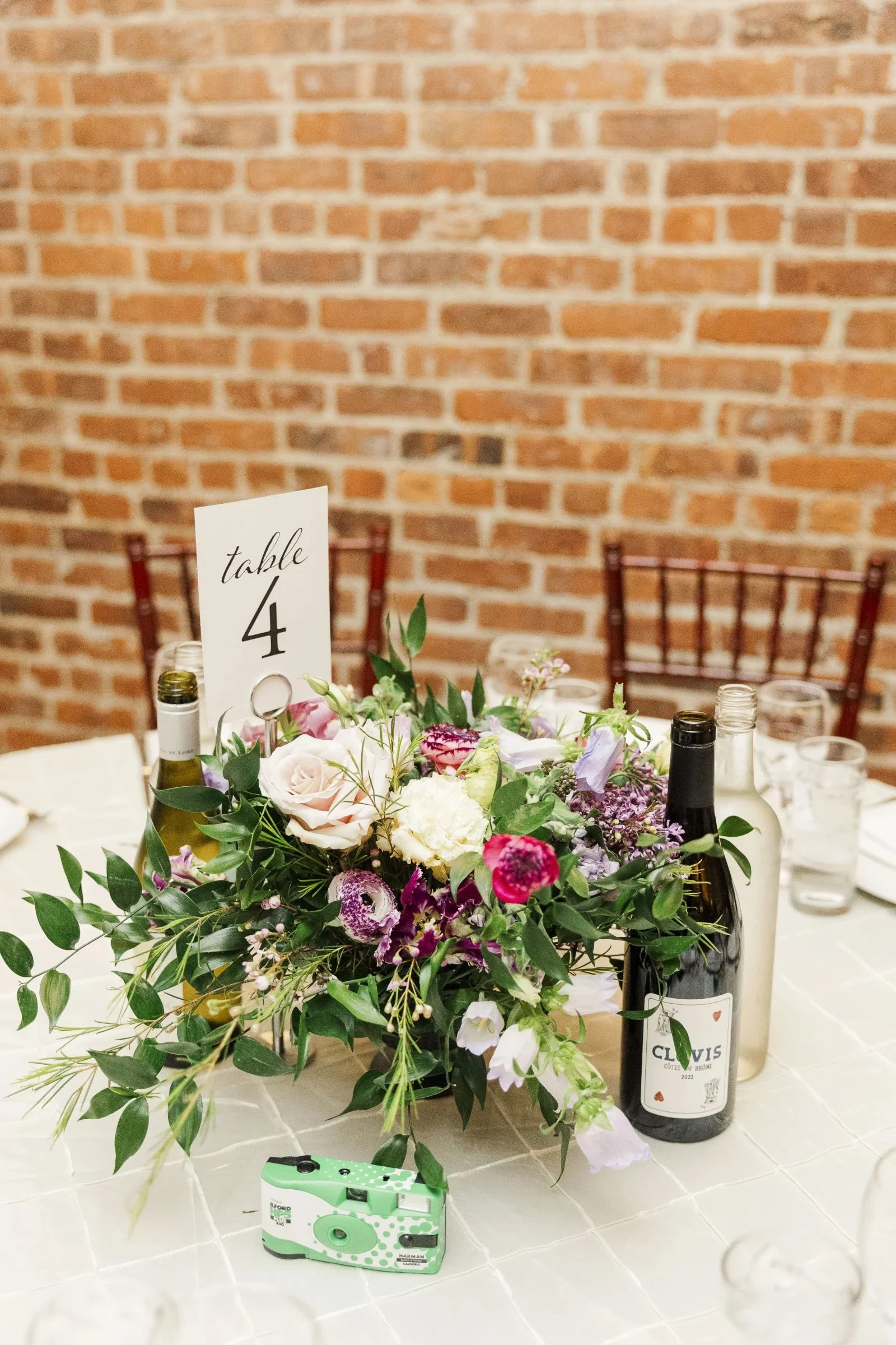 Flower arrangement with pink, purple, and white flowers, surrounded by greenery, on a table with wine bottles, glasses, and a table number sign reading 'table 4,' set against a brick wall background.