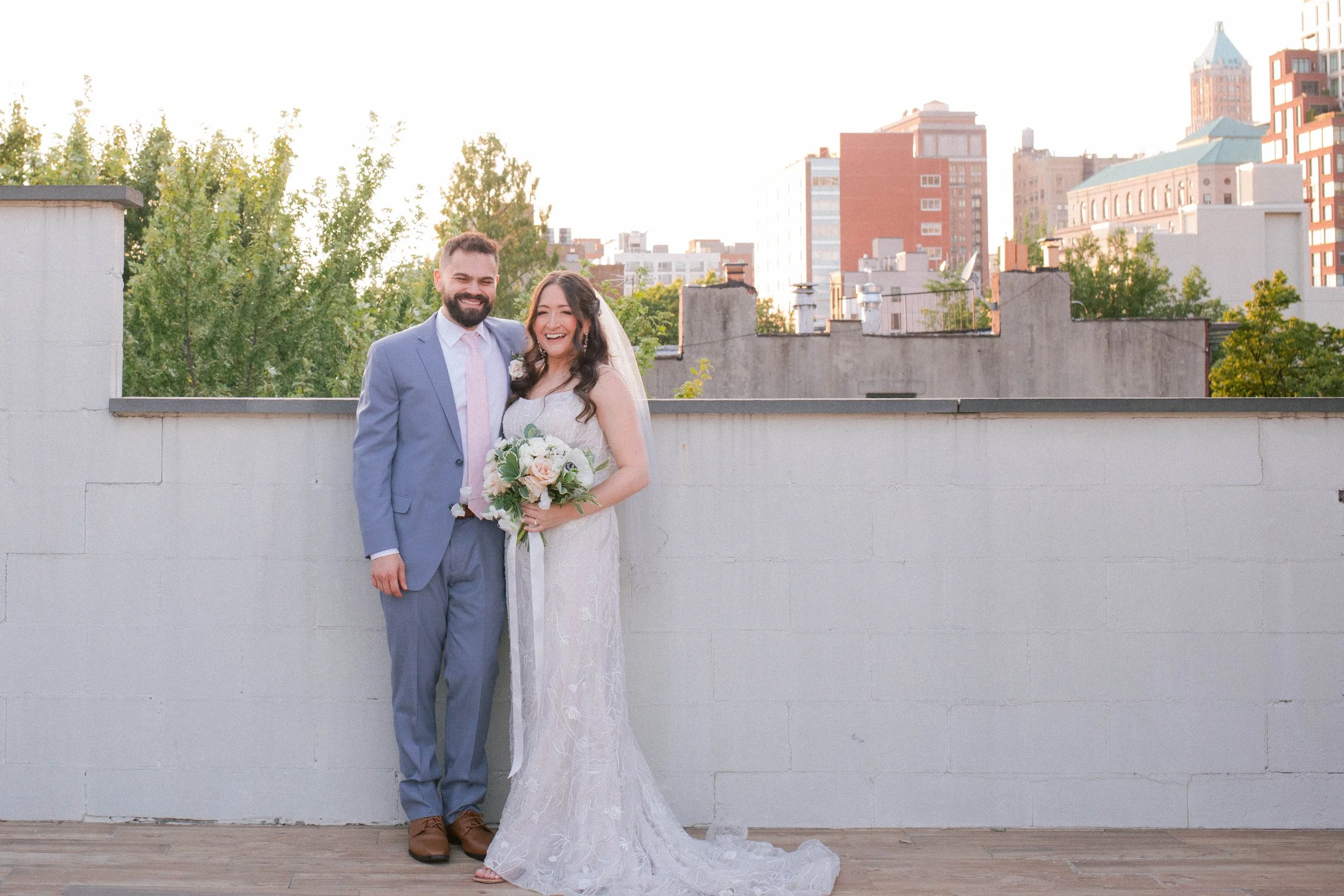A newlywed couple standing on a rooftop terrace with a cityscape in the background, smiling and holding a bouquet of flowers.