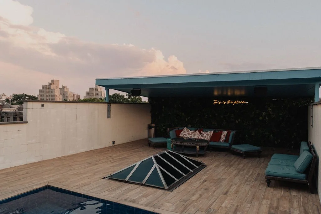 Rooftop patio with seating area, lounge chairs, and a glass pyramid skylight, overlooking city skyline at sunset.