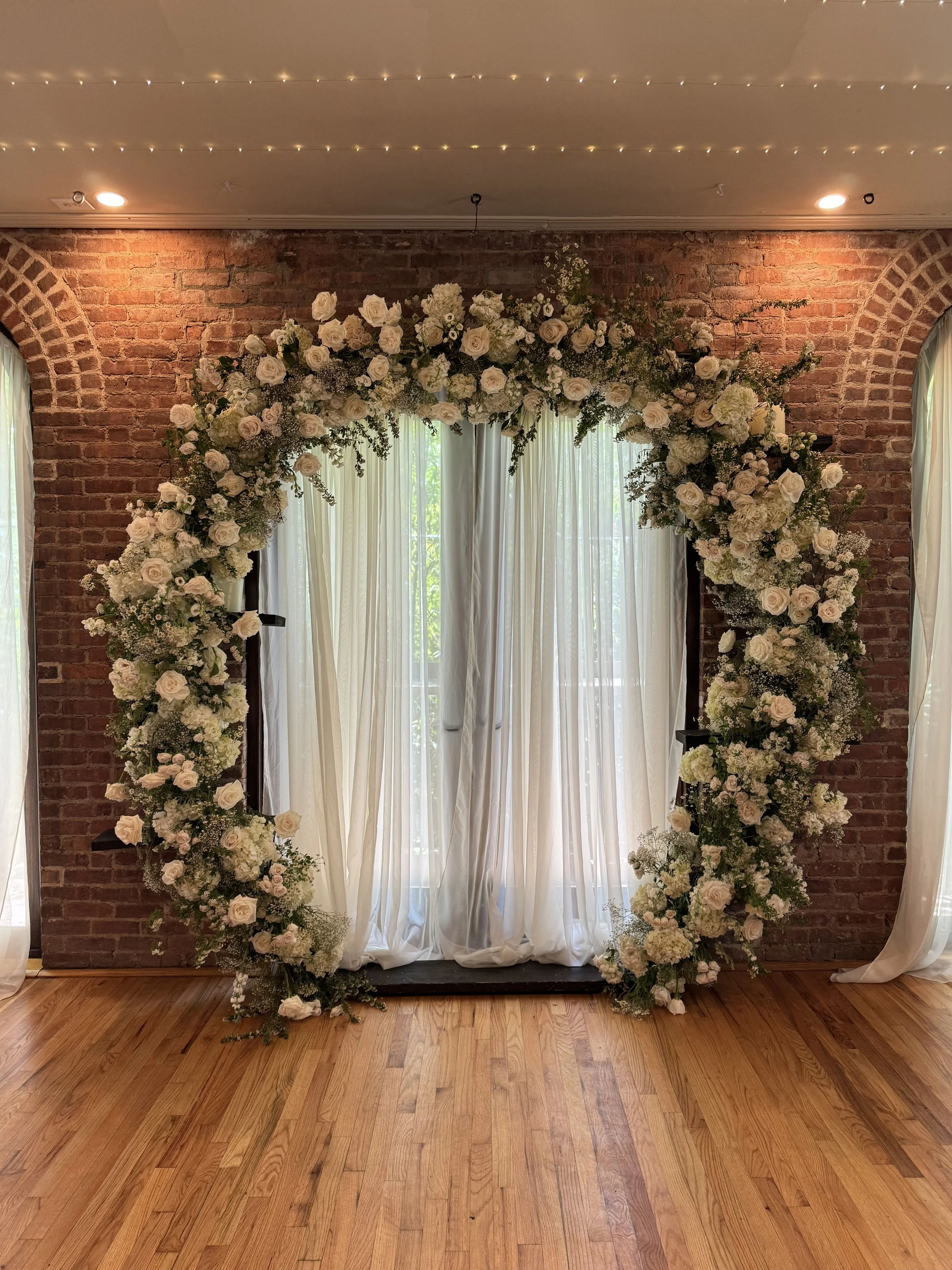 A floral arch decorated with white roses, hydrangeas, and greenery set against a brick wall with hanging fairy lights, white curtains, and a wooden floor.