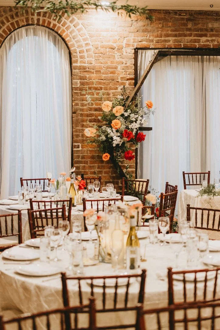 A decorated event space with round tables set for a meal, including glasses, plates, and bottles. The room has brick walls, tall windows with sheer curtains, and a floral arrangement on a diagonal wooden beam against the brick wall, featuring peach, 