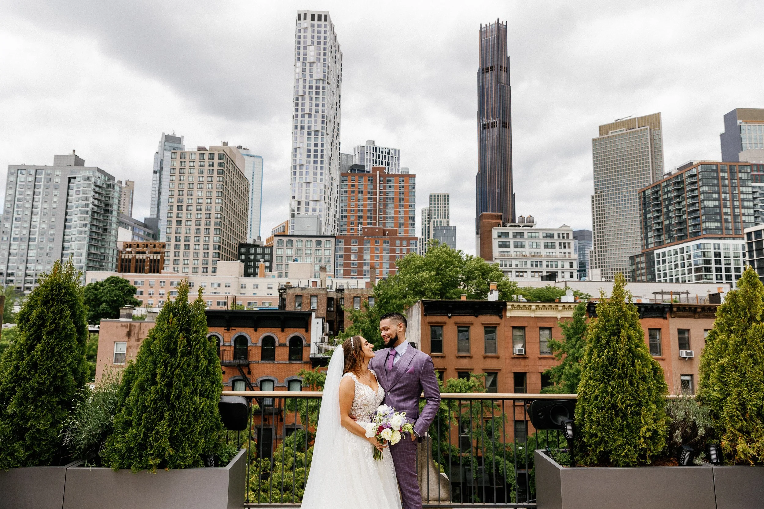 Wedding couple standing in an urban garden with city skyscrapers in the background, trees and greenery in the foreground.