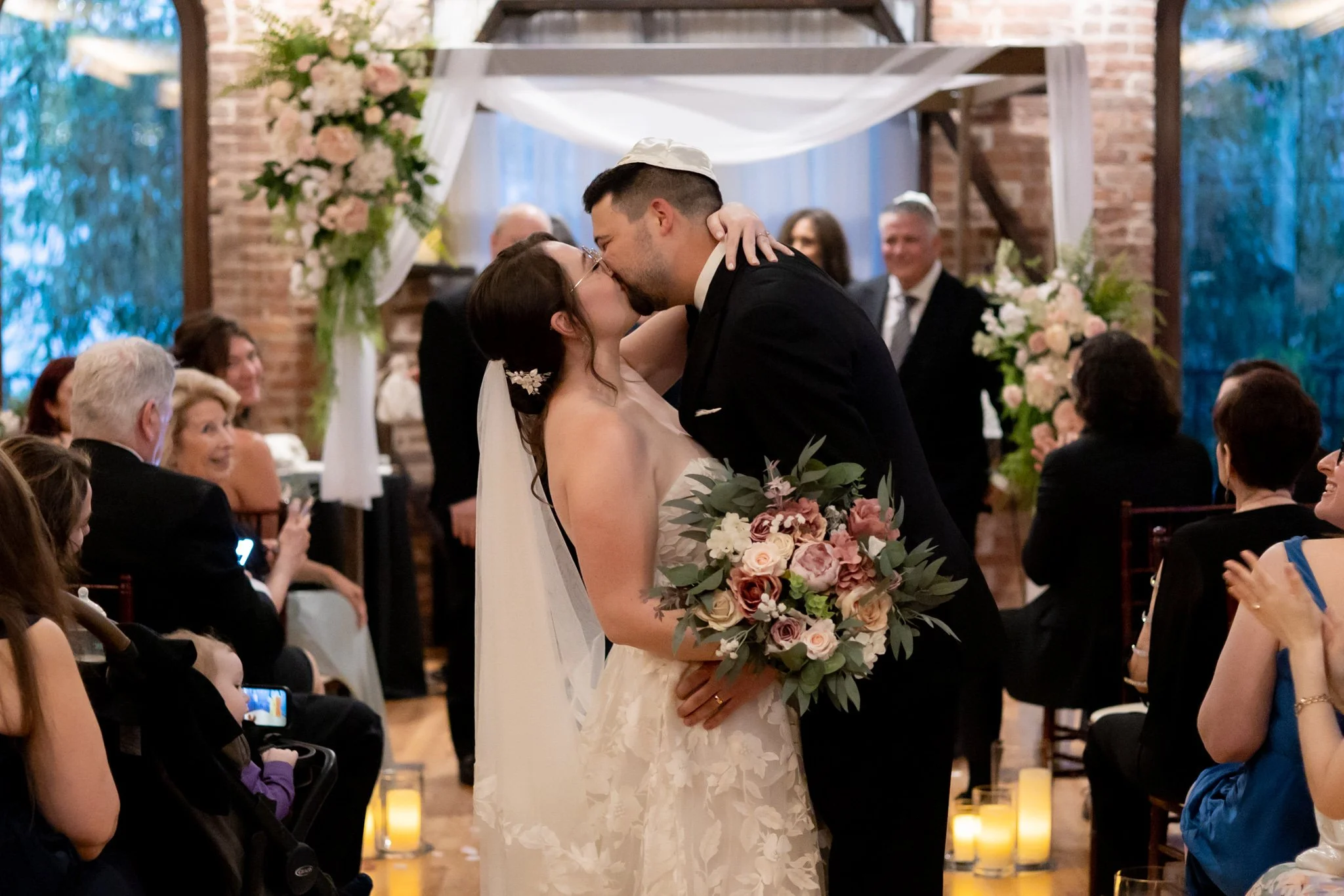 A couple sharing a kiss during their wedding ceremony. The bride is dressed in a white lace dress, holding a bouquet of pink and white flowers. The groom is wearing a black suit and a white cap. Guests are seated around them, some taking photos, with