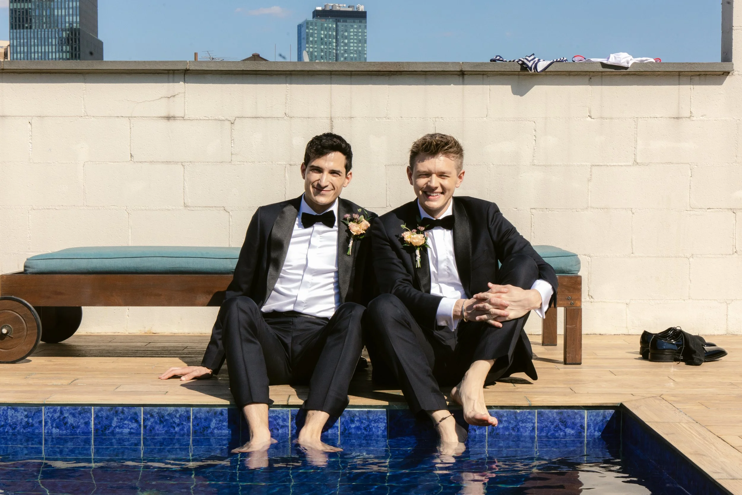 Two men in tuxedos with flower boutonnieres sitting on a poolside deck with their feet in the water, smiling at the camera, against a city skyline background.