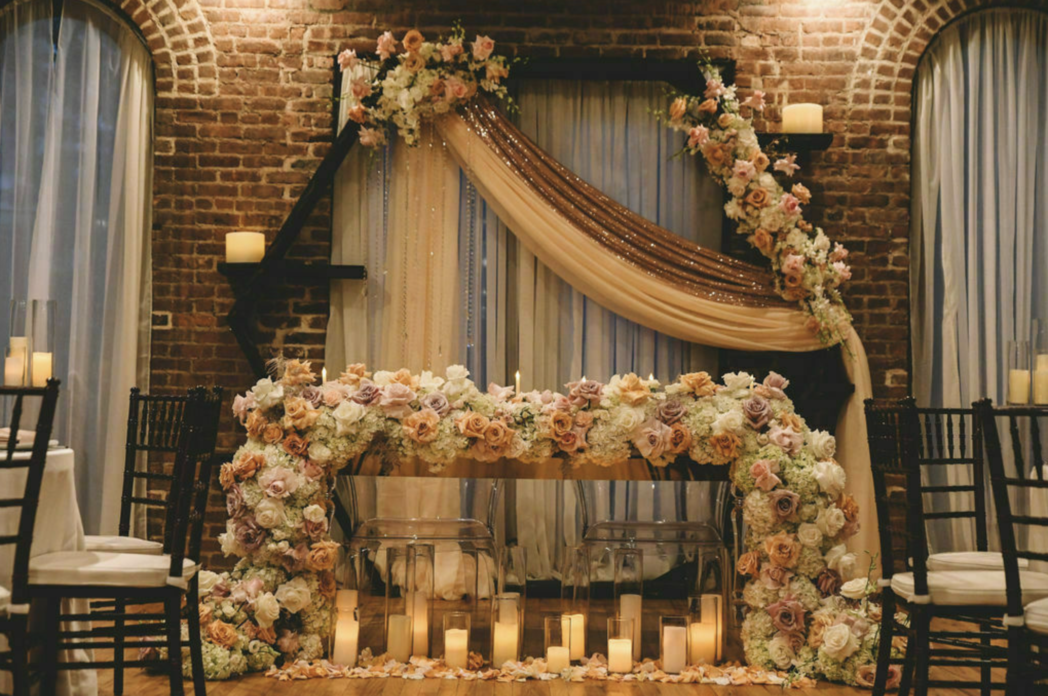 Elegant wedding ceremony backdrop with cascading floral arrangements of roses and peonies, draped beige and copper curtains, and candles on a brick wall setting.