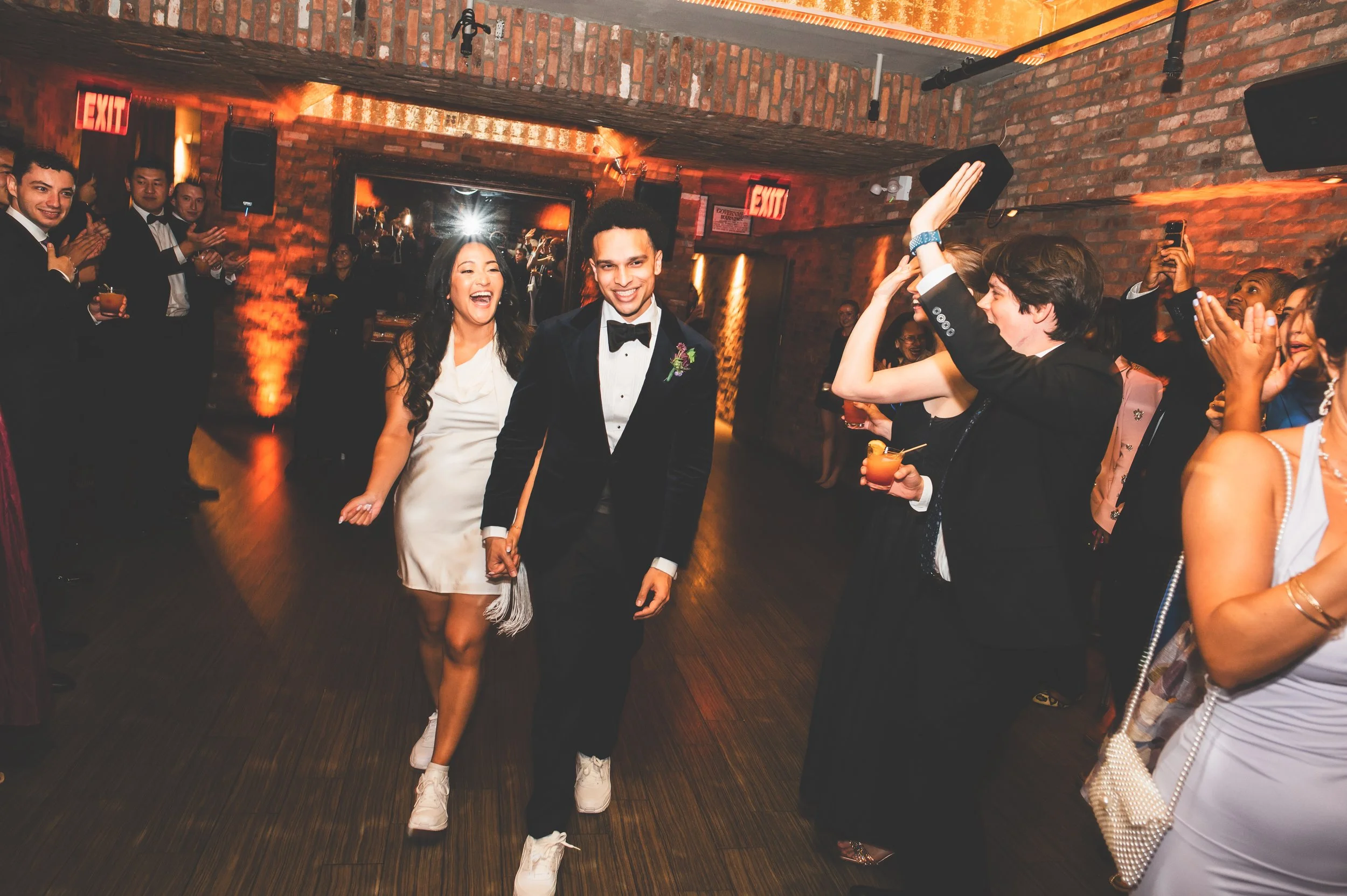 A bride and groom holding hands and dancing at their wedding reception, surrounded by friends and family in a brick-walled venue.