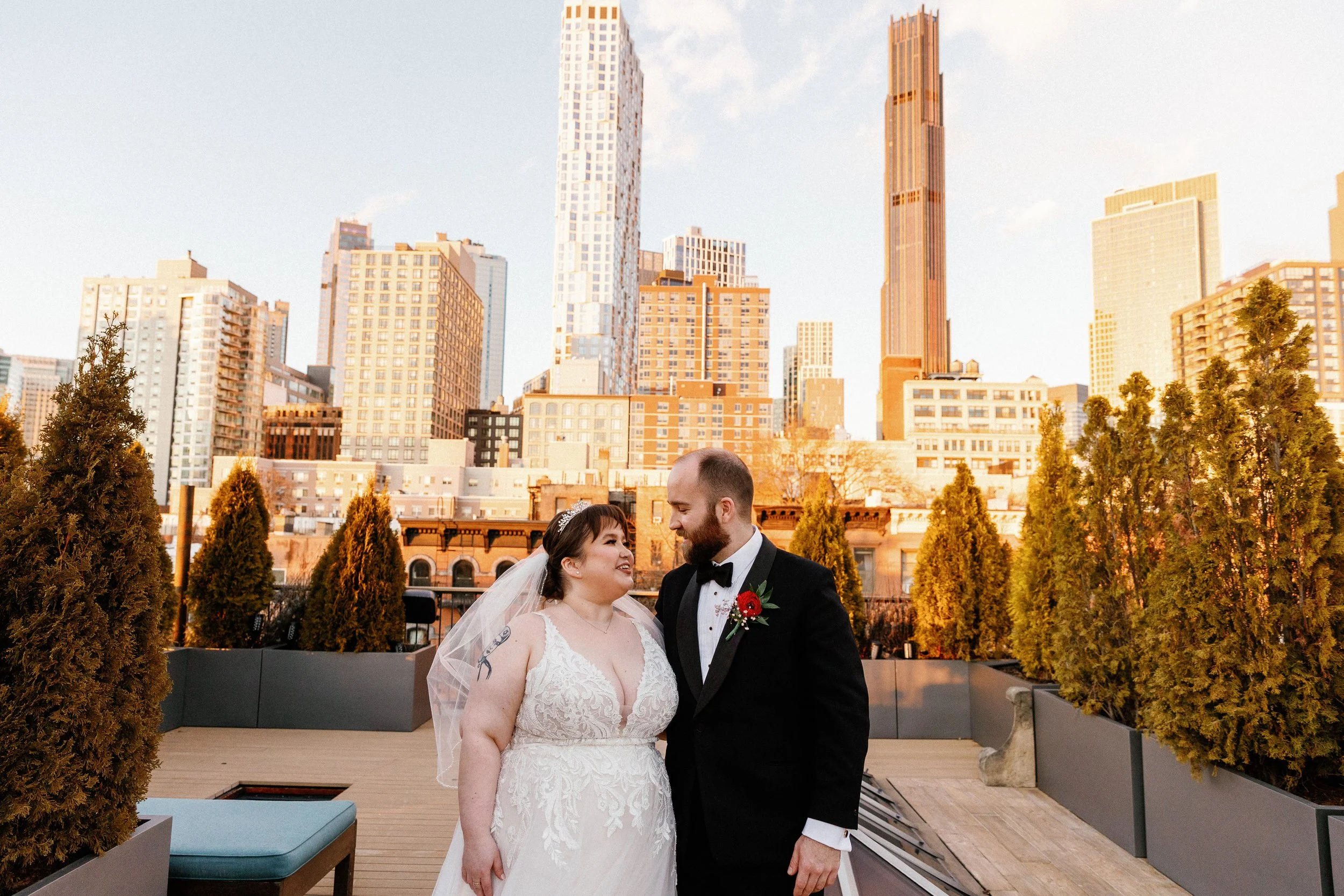 A newlywed couple stands close together on a rooftop in front of a city skyline with tall buildings, trees, and blue sky during sunset.