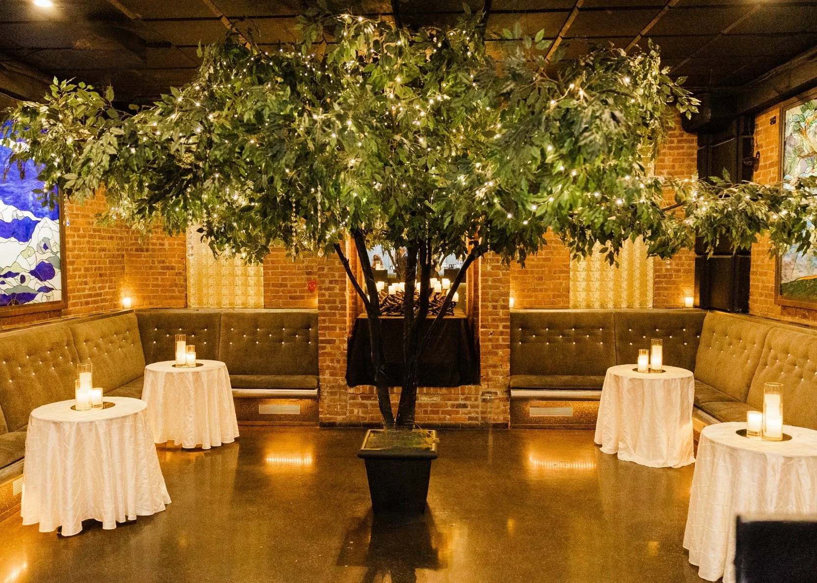 Indoor setting with a large potted tree decorated with string lights, surrounding corner booths with tufted upholstery, and small round tables with white tablecloths and candles, brick wall and stained glass windows in the background.
