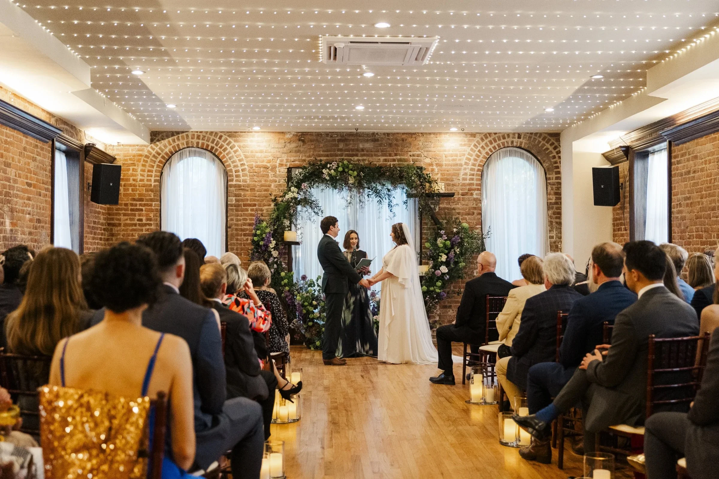 A wedding ceremony taking place in a brick-walled venue with soft lighting, where a couple is exchanging vows in front of an officiant, surrounded by seated guests.