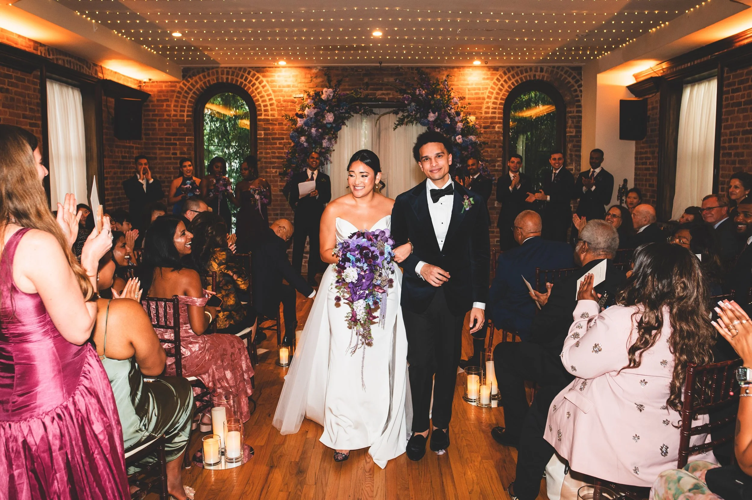 A bride and groom walking down the aisle at their wedding ceremony, surrounded by seated guests in a decorated indoor venue with brick walls, arched windows, floral arrangements, and candles.