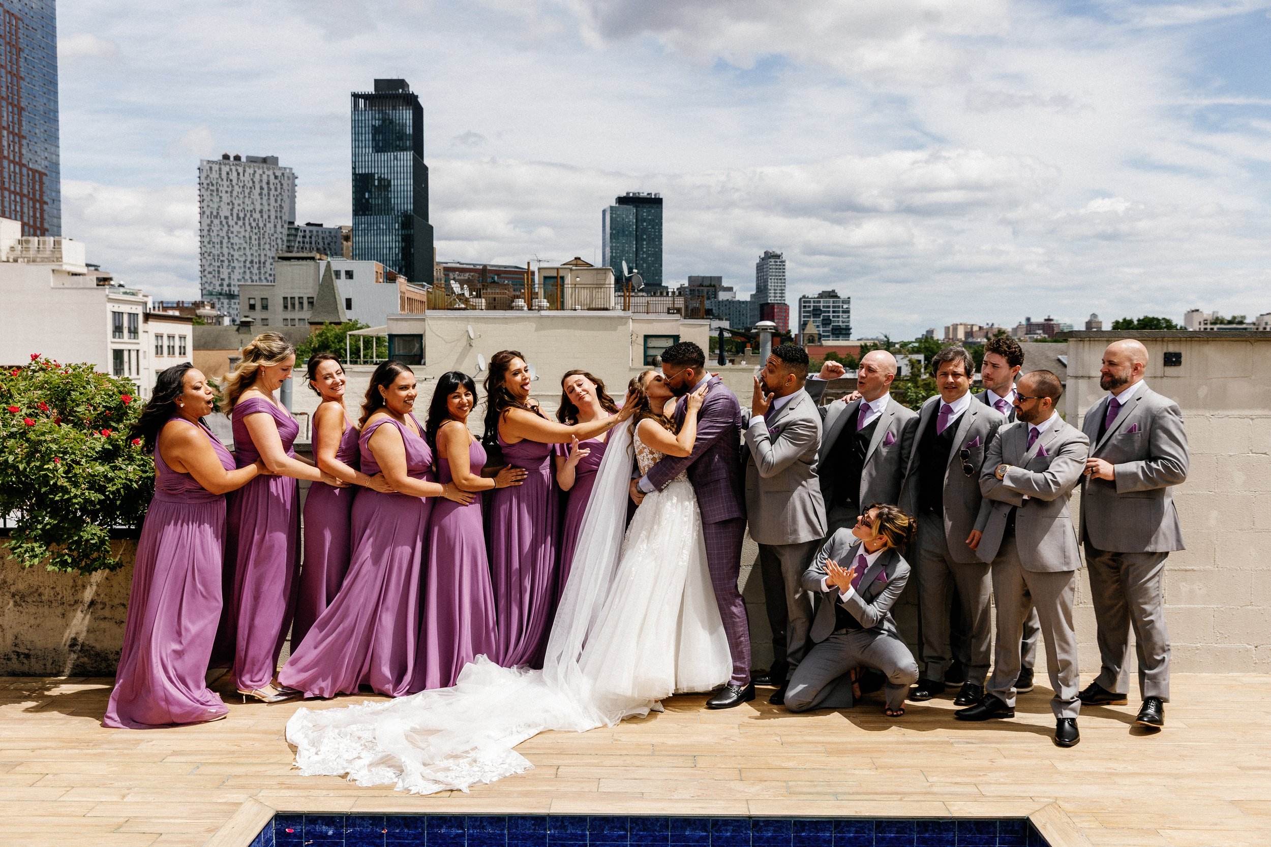 A group of women in purple dresses and men in gray suits are celebrating a wedding on a rooftop with a city skyline in the background, including tall modern buildings and a cloudy sky.
