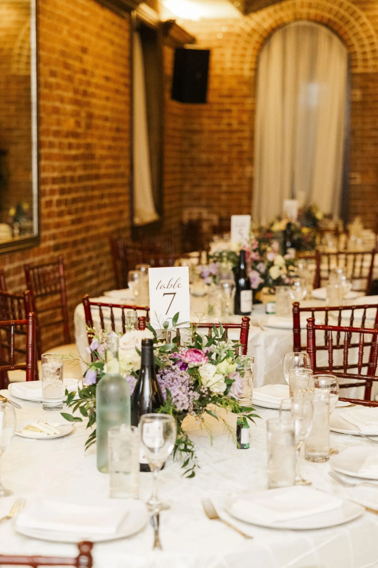 A wedding reception table decorated with a floral centerpiece, wine bottles, and table settings, with a brick wall and window with curtains in the background.