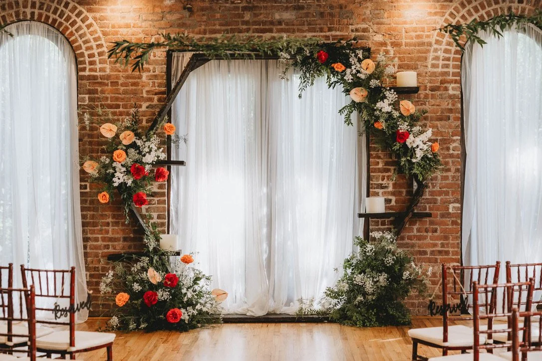 Wedding or event backdrop with a floral arch and chairs in front, set against a brick wall with arched windows and white curtains.