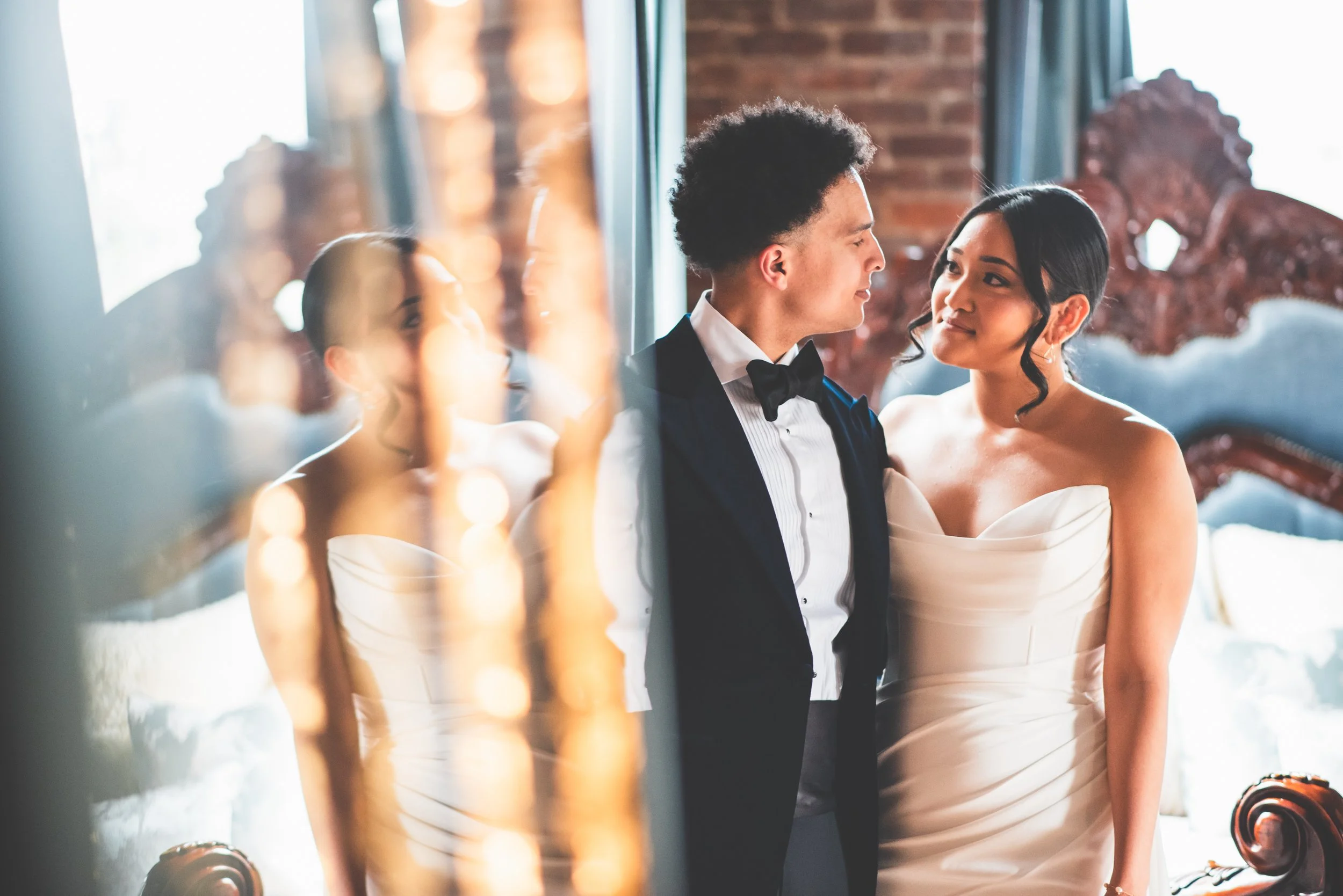 A bride and groom look at each other in a close pose inside a room, with warm lighting and a brick wall background. The photo is partially reflected in a mirror with lights around it.