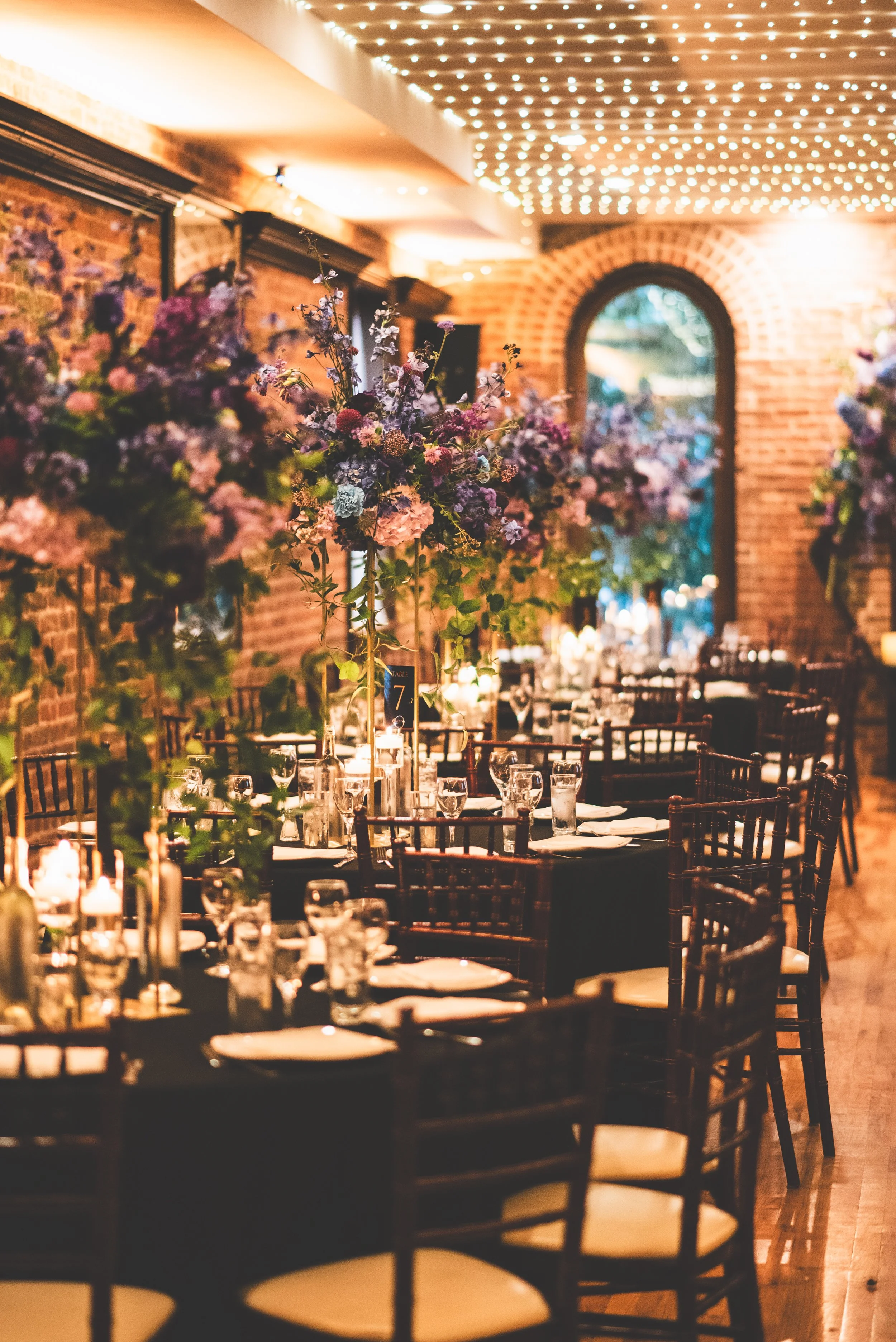 Elegant indoor dining area with flower centerpieces, neatly set tables, exposed brick walls, and string lights on the ceiling for ambiance.