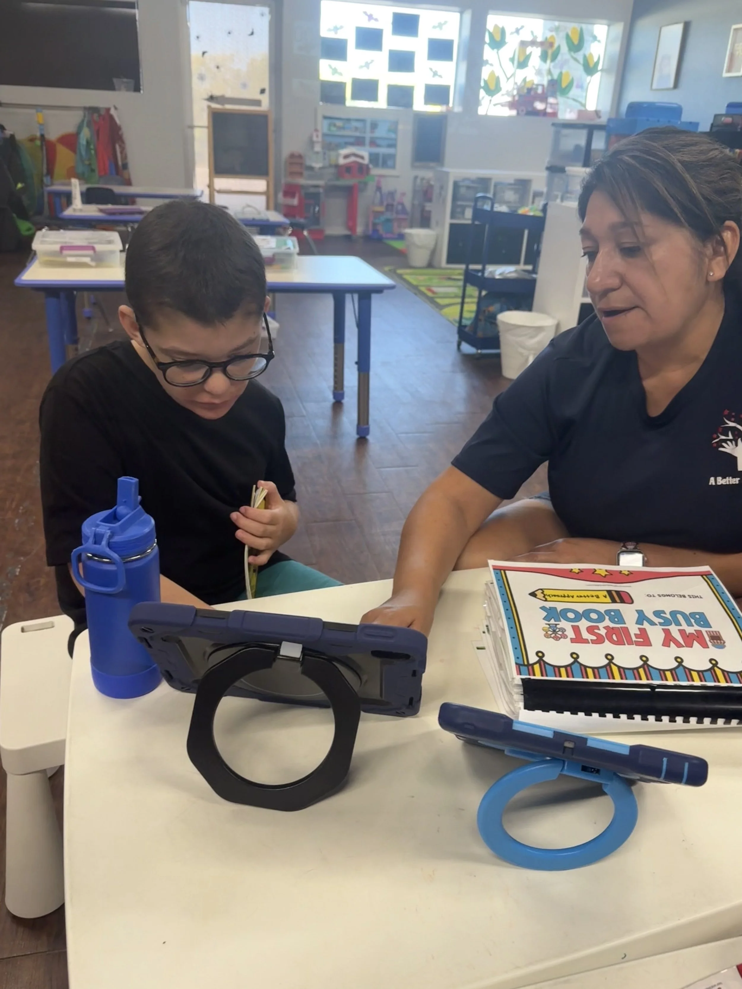 A young boy with glasses and a woman sitting at a table in a classroom, looking at a tablet computer on a stand. The table also has a blue water bottle, a binder labeled 'My First Busy Book,' and a blue pen. The classroom has colorful educational decor and windows in the background.
