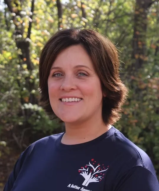 A woman with short brown hair smiling outdoors with trees in the background, wearing a navy blue shirt with a logo and text.