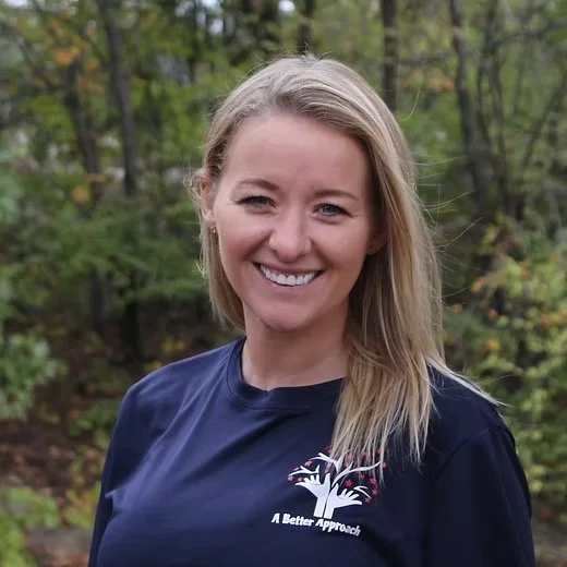 Woman smiling outdoors in a wooded area, wearing a navy blue t-shirt with a logo of a tree and the text "A Better Approach."