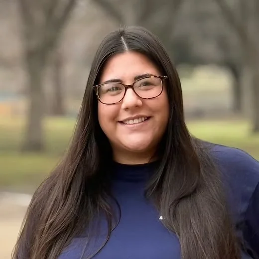 A smiling woman with long dark hair, wearing glasses and a navy blue shirt, standing outdoors in a park with blurred trees in the background.