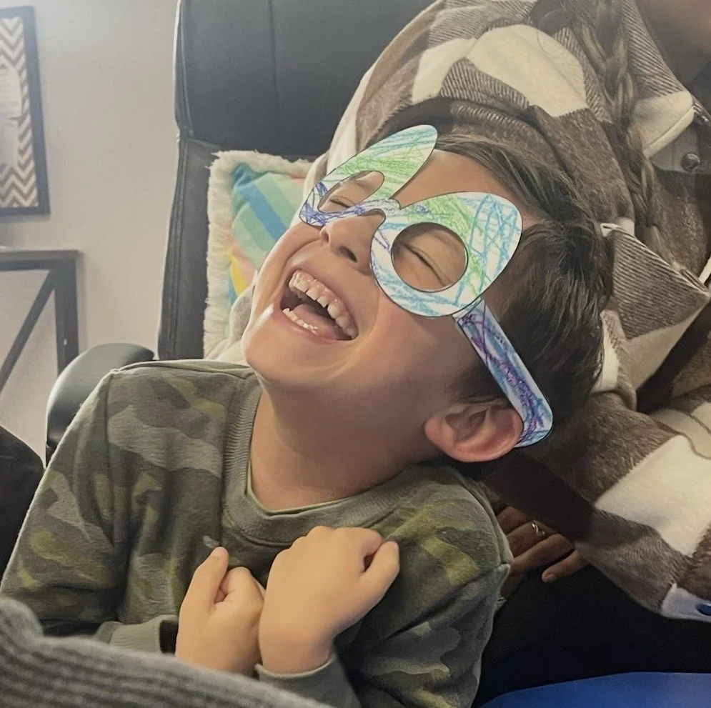 A young boy wearing colorful, oversized glasses with blue, green, and purple crayon drawings, laughing joyfully with hands clenched in front of his chest, sitting on a couch with colorful pillows behind him.