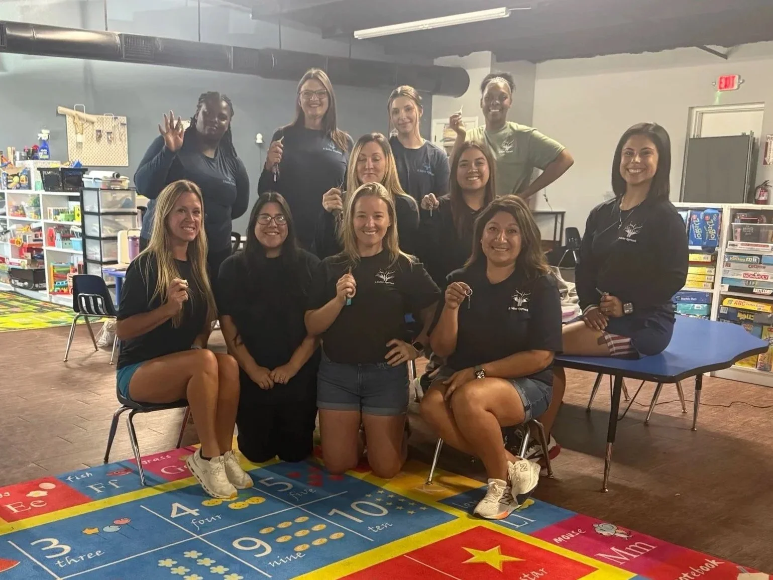 Group of women in casual attire sitting and standing around a colorful number and letter rug in a classroom or play area, smiling at the camera.