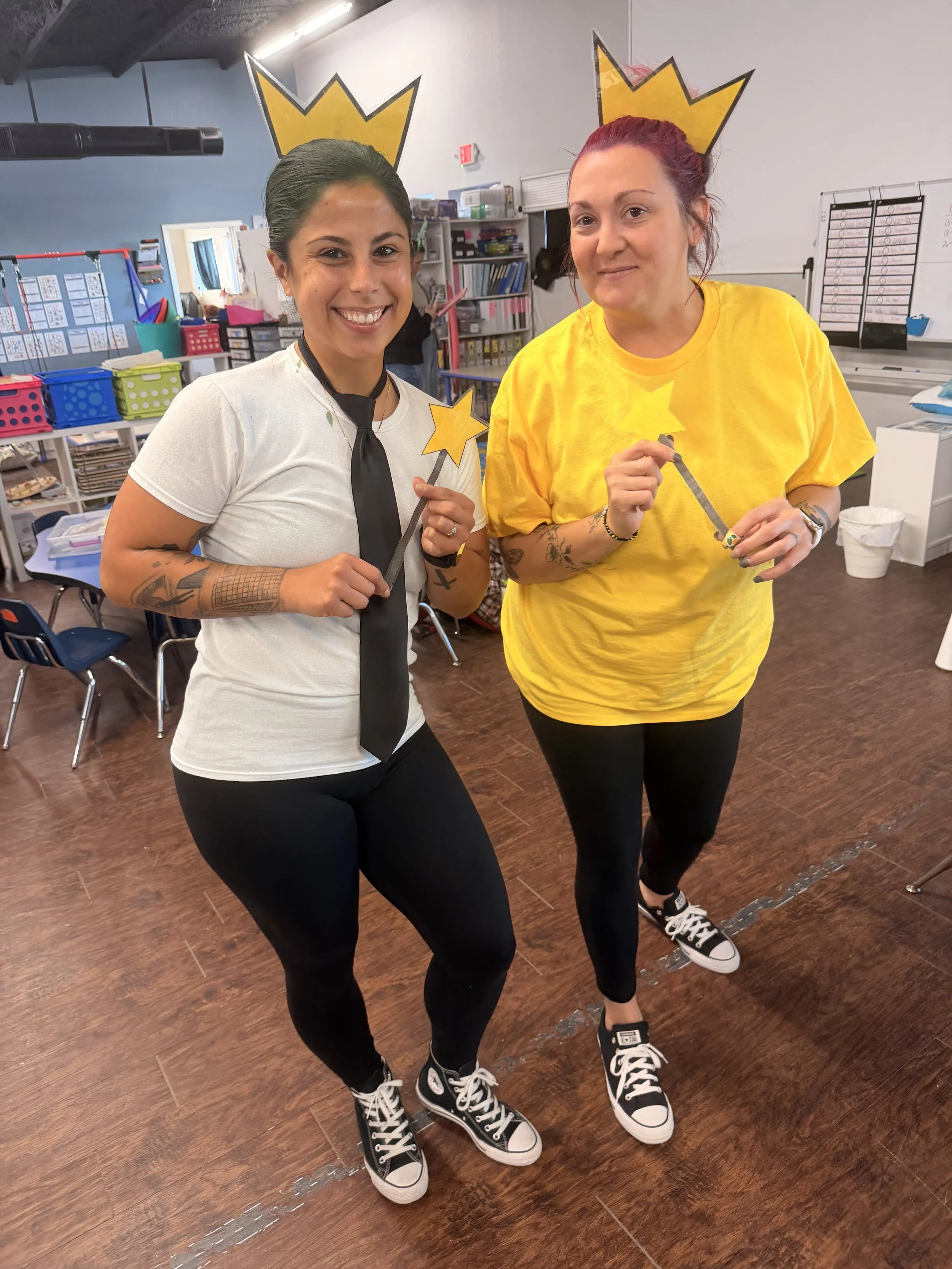 Two women wearing cardboard crown and wand props, smiling in a classroom decorated with crown illustrations, tables, chairs, and educational supplies.