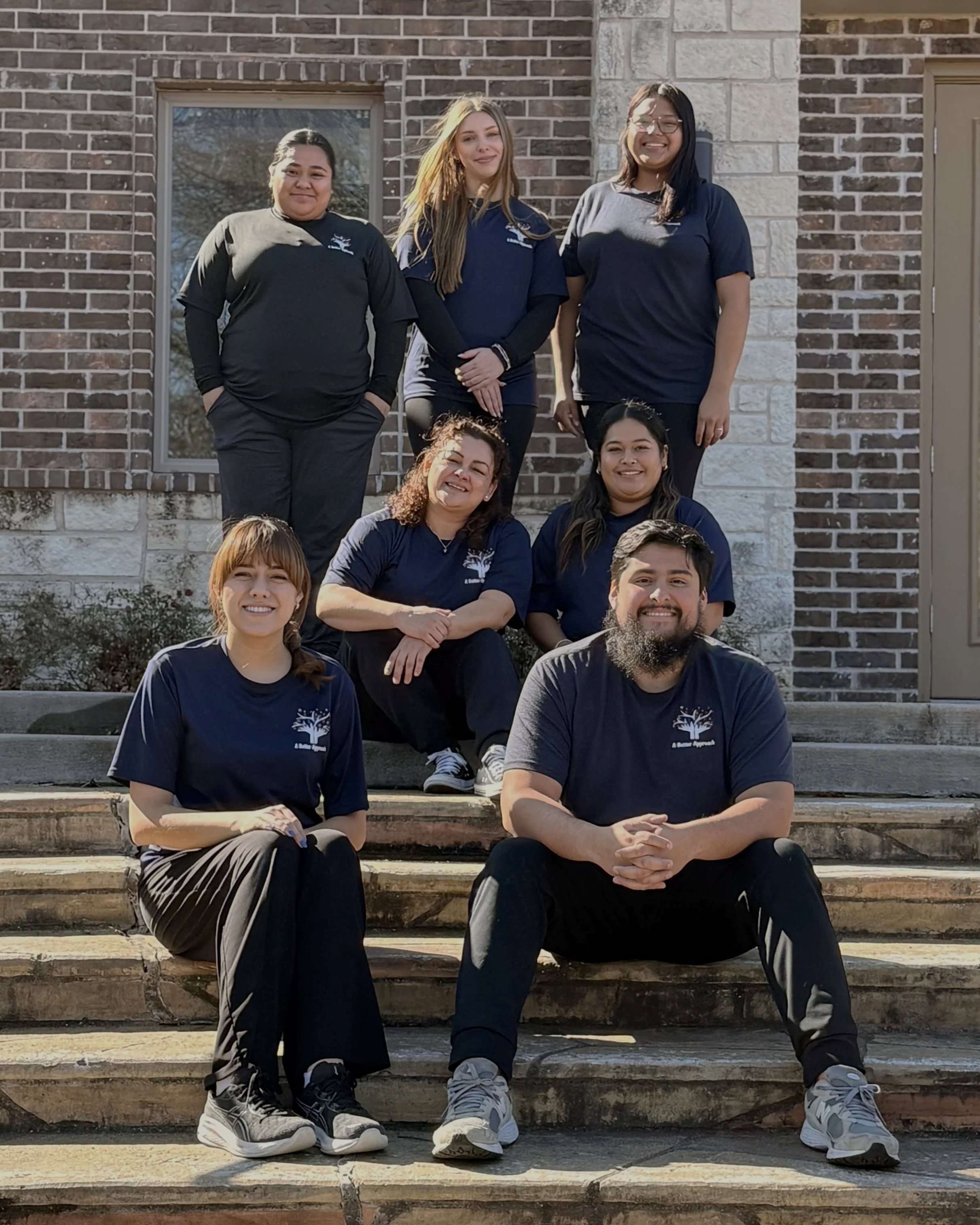 Group of seven diverse people posing on steps in front of a brick building, all wearing matching navy blue and black shirts with a tree logo.