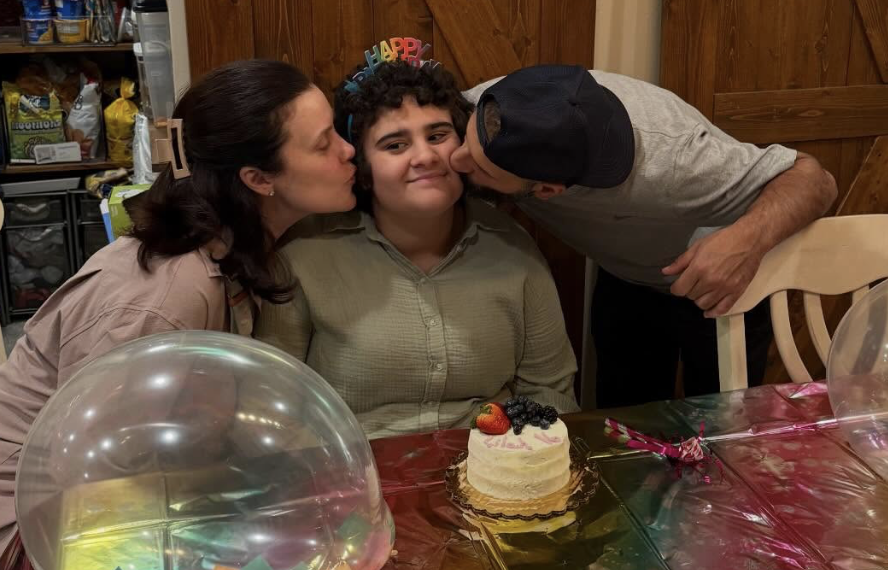 A mom and dad kissing their daughter sitting in front of a birthday cake