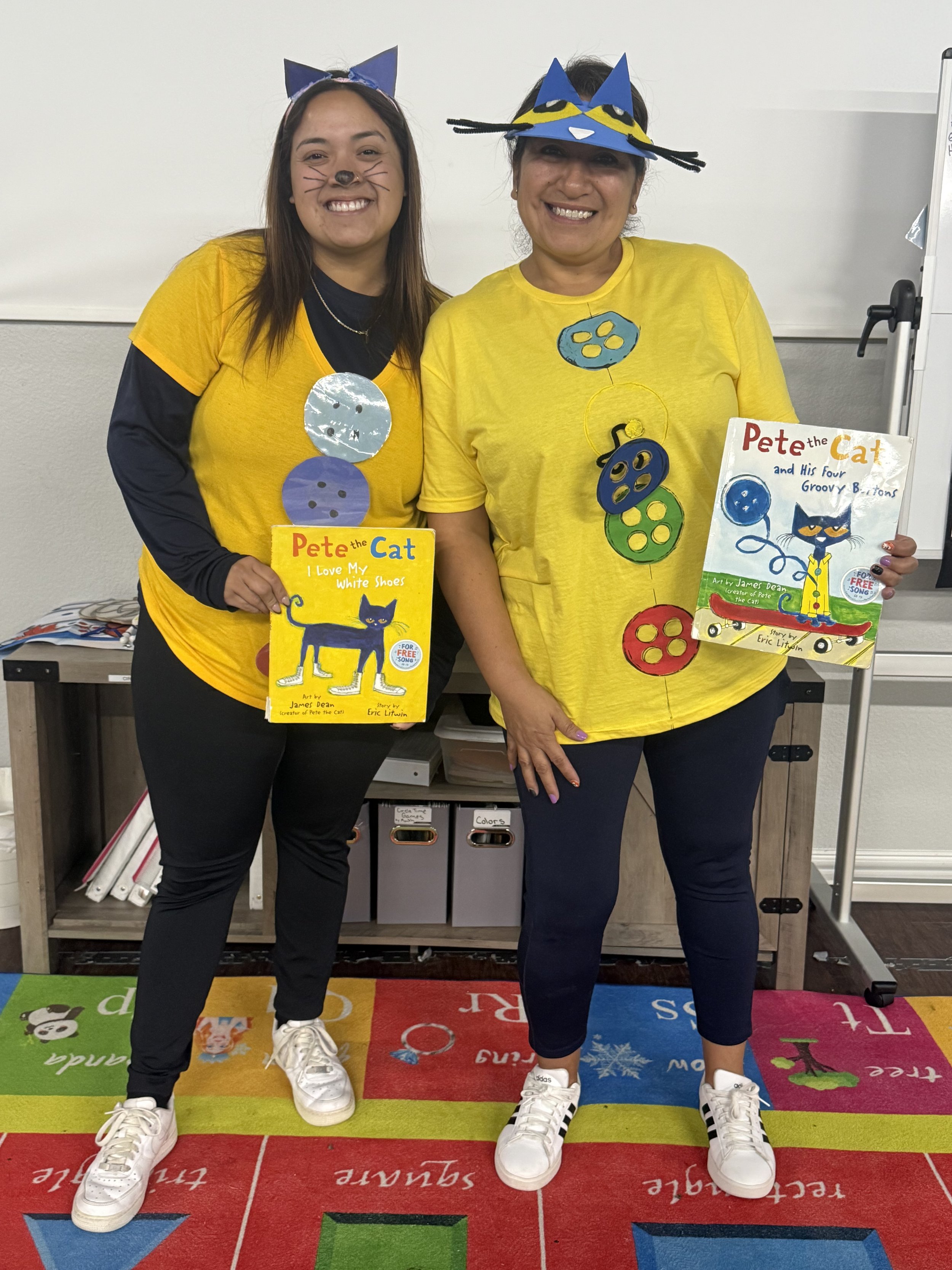 Two women wearing yellow t-shirts with Pete the Cat themed decorations, standing indoors on a colorful educational rug, each holding a Pete the Cat book.
