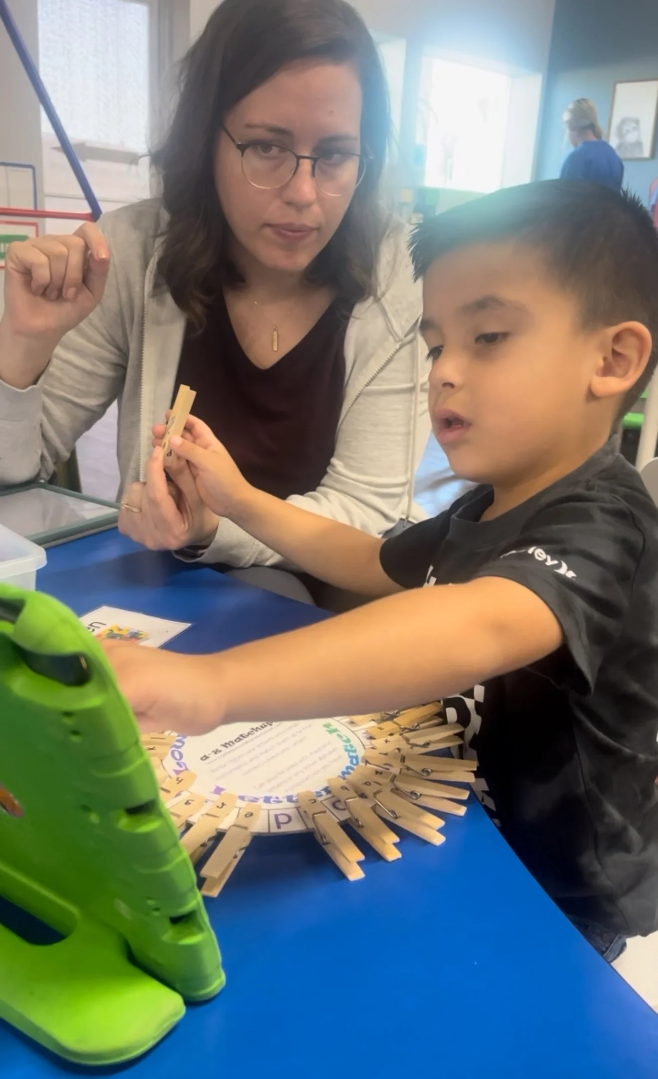 A woman and a young boy sitting at a table, interacting with a matching game using clothespins and cards. The woman is looking at the boy, who is attaching a clothespin to a card on the table.