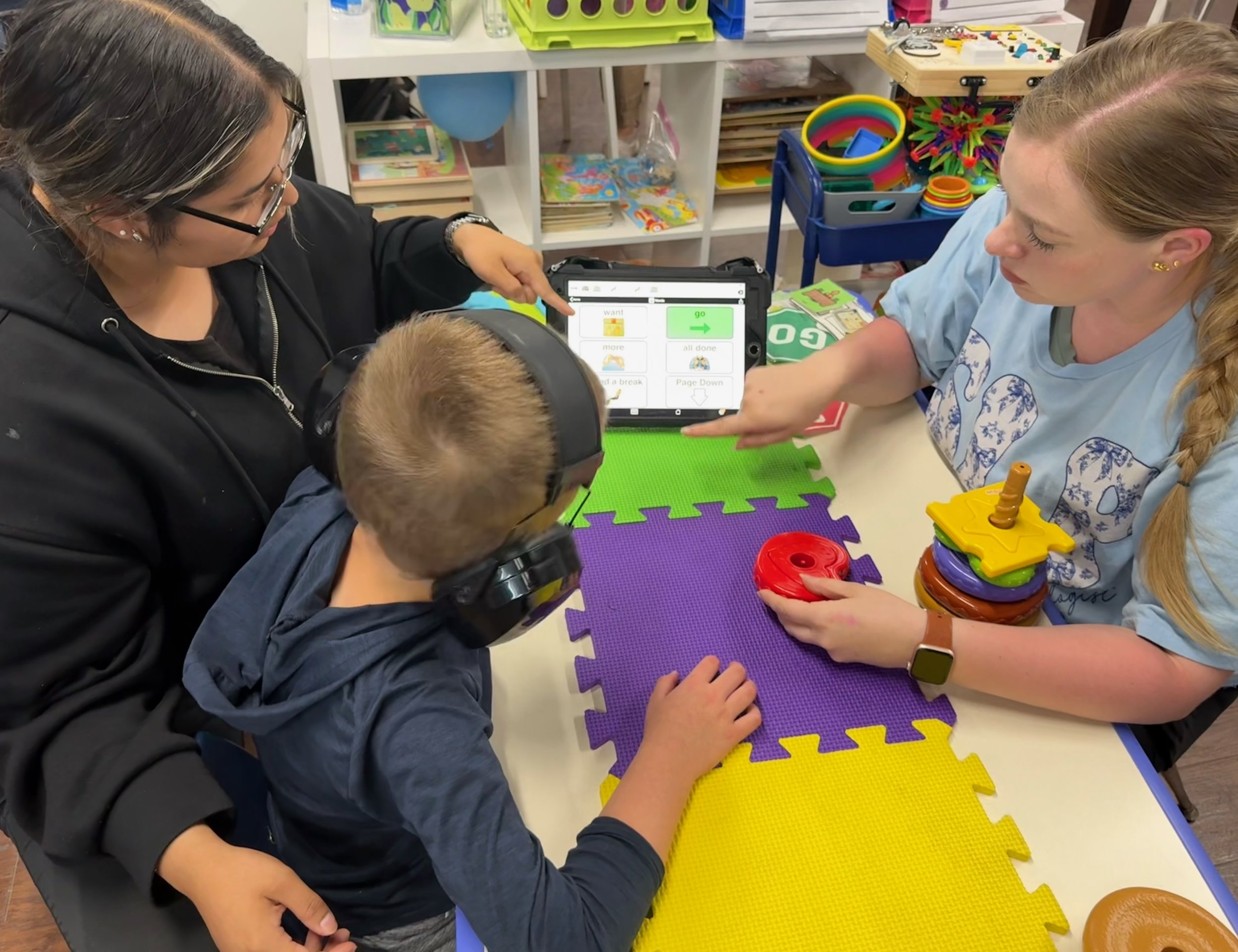 Teacher and children engaged in a learning activity at a table with colorful foam mats, in a classroom setting with educational toys and books in the background.