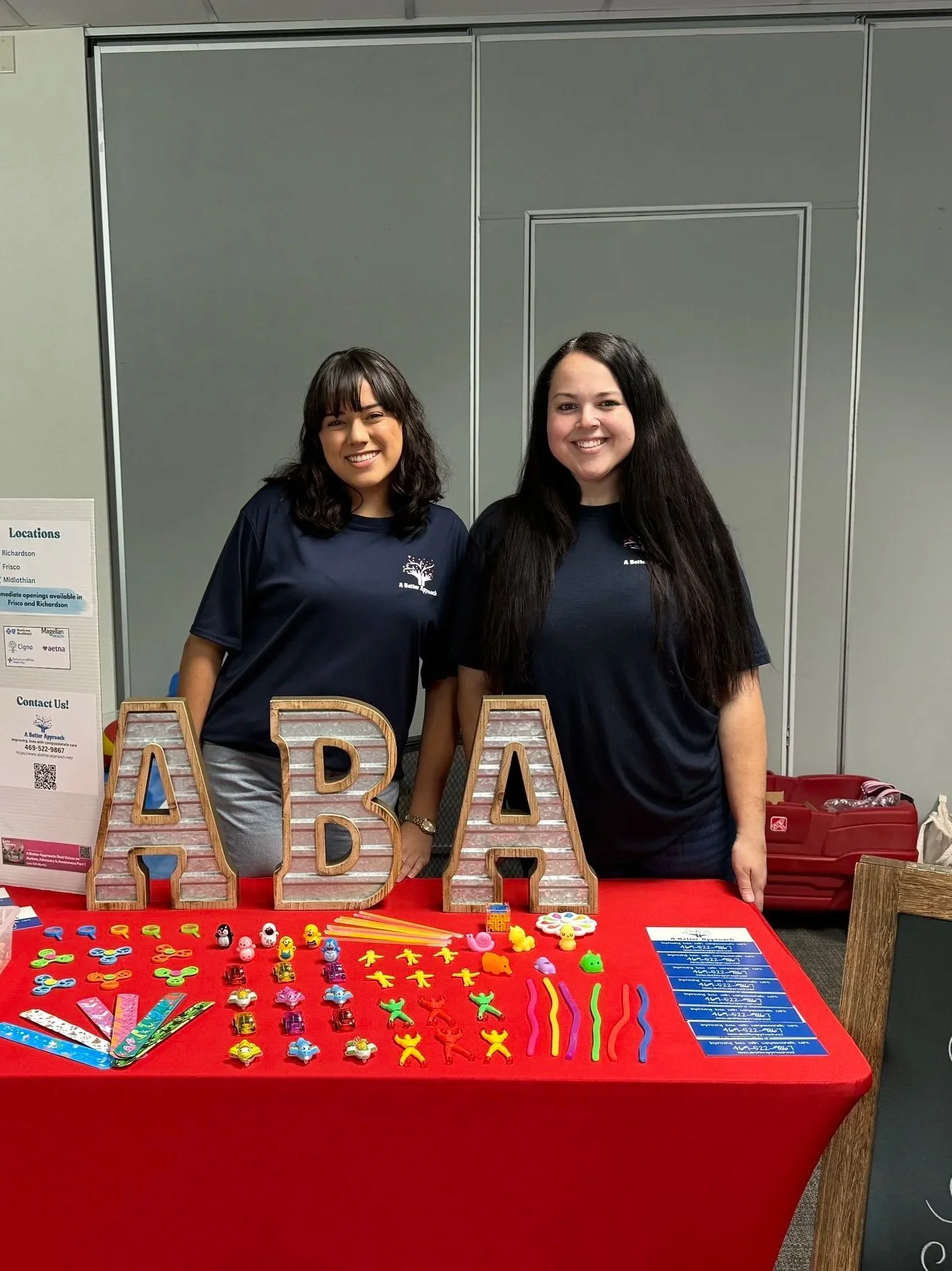 Two women standing behind a table decorated with colorful toys, with large wooden letters spelling 'ABA' in front. Both women are wearing dark blue shirts.