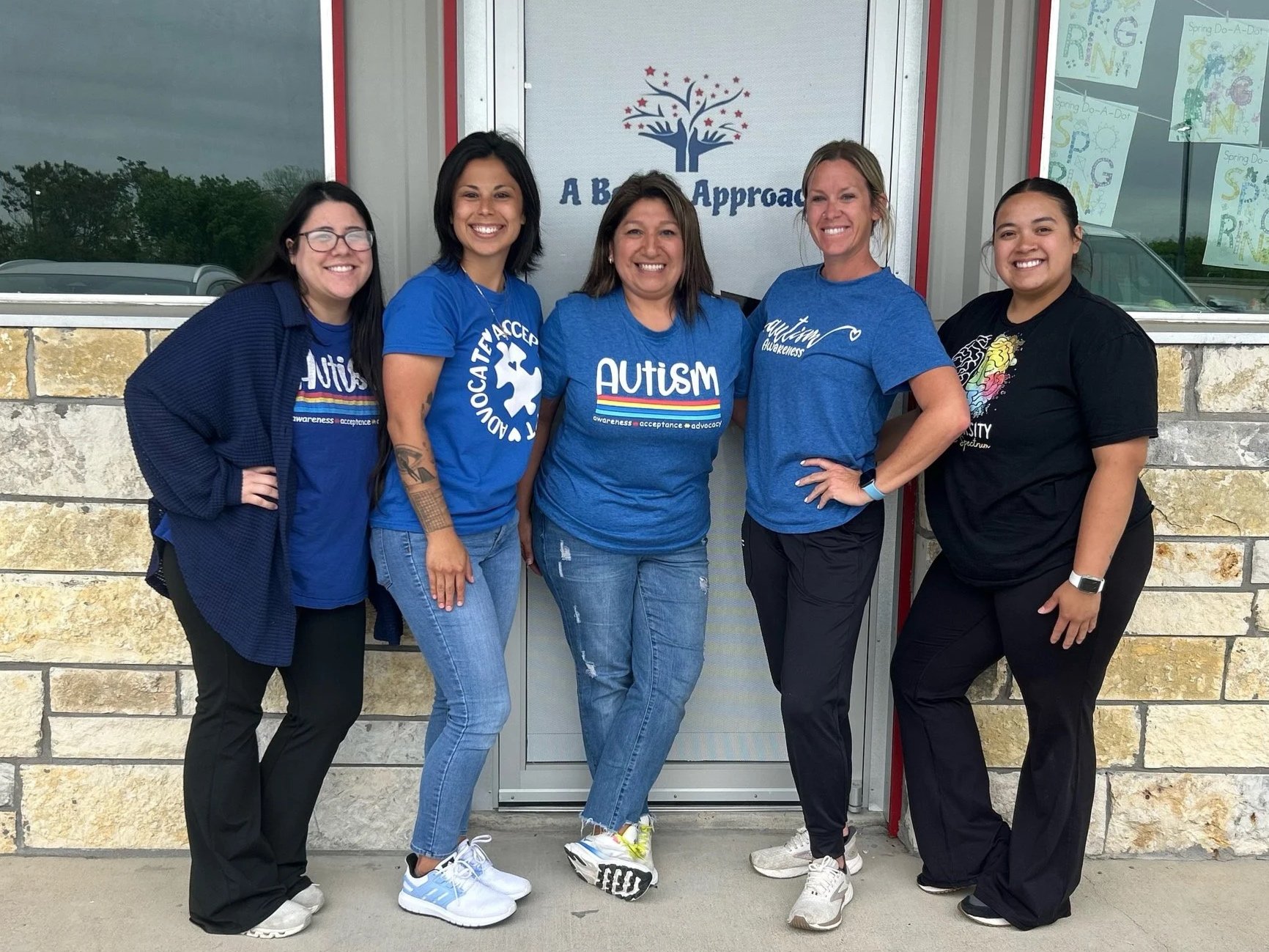 Five women standing outside in front of a door with an 'Autism' sign. They are smiling at the camera, dressed casually, with some wearing blue shirts supporting autism awareness.