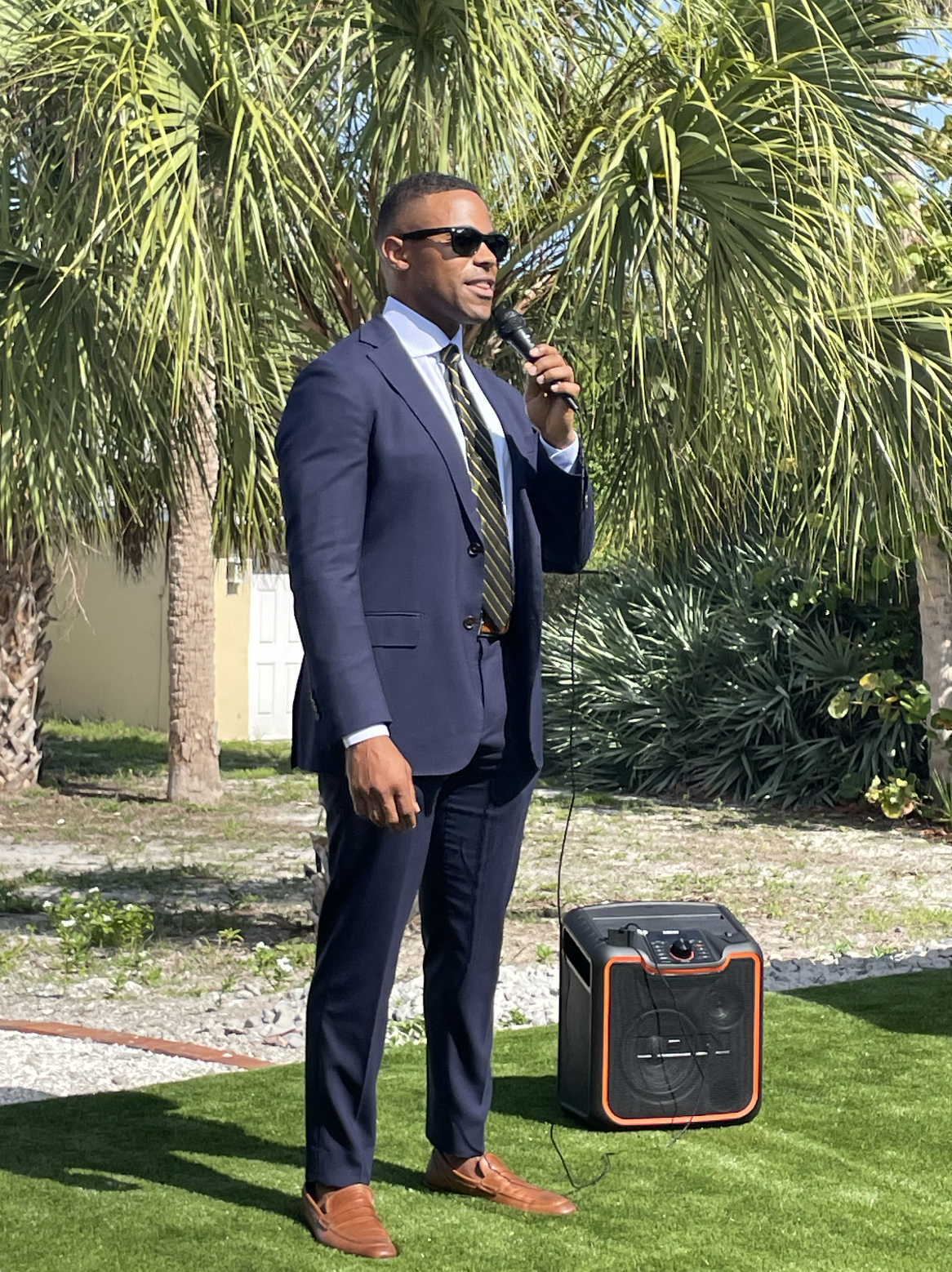 A man in a navy suit, sunglasses, and tie, holding a microphone, standing outdoors near green palm trees and a portable speaker.