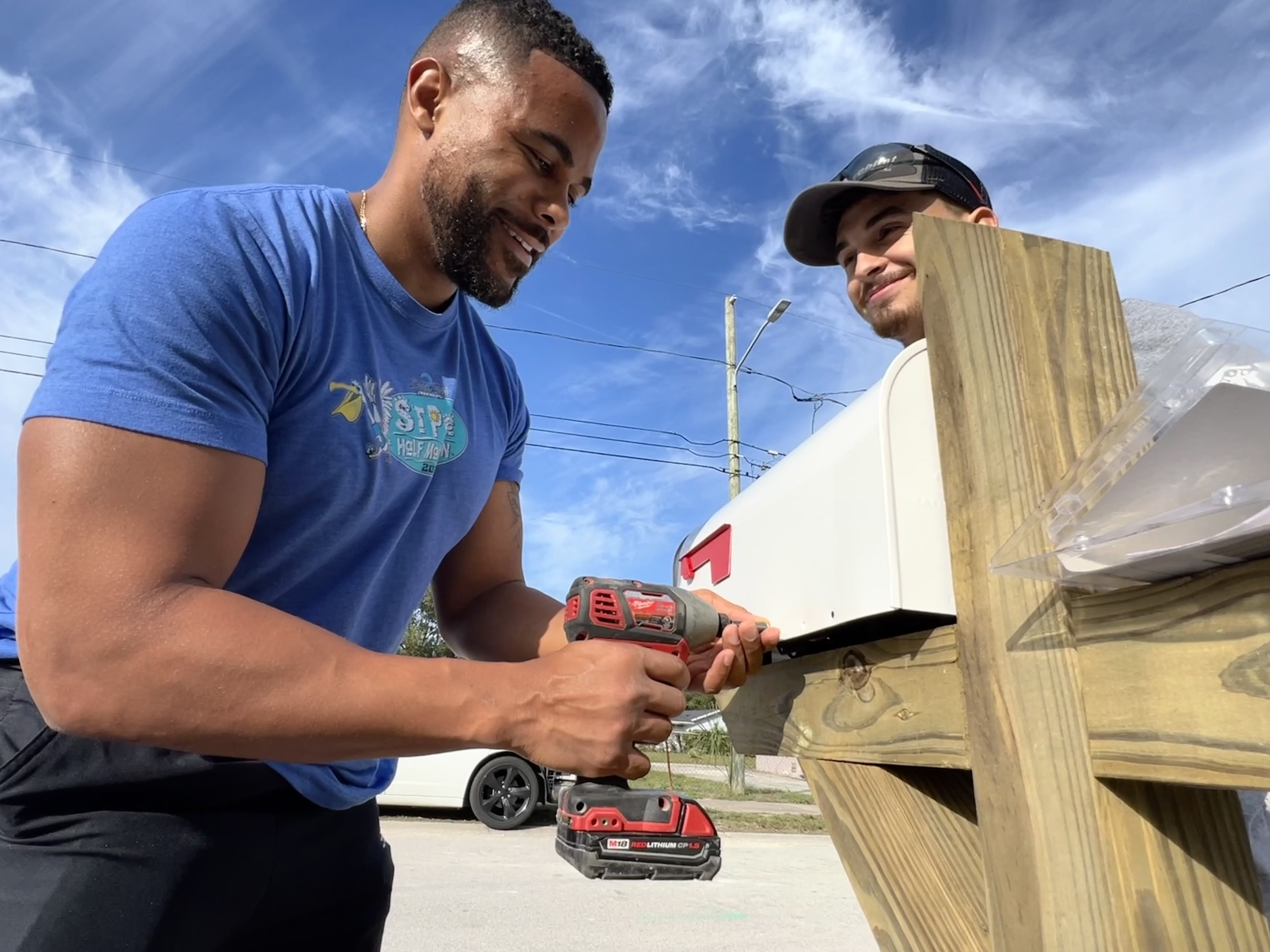 Two men working outdoors on a wooden project, one using a cordless drill, smiling under a clear blue sky.