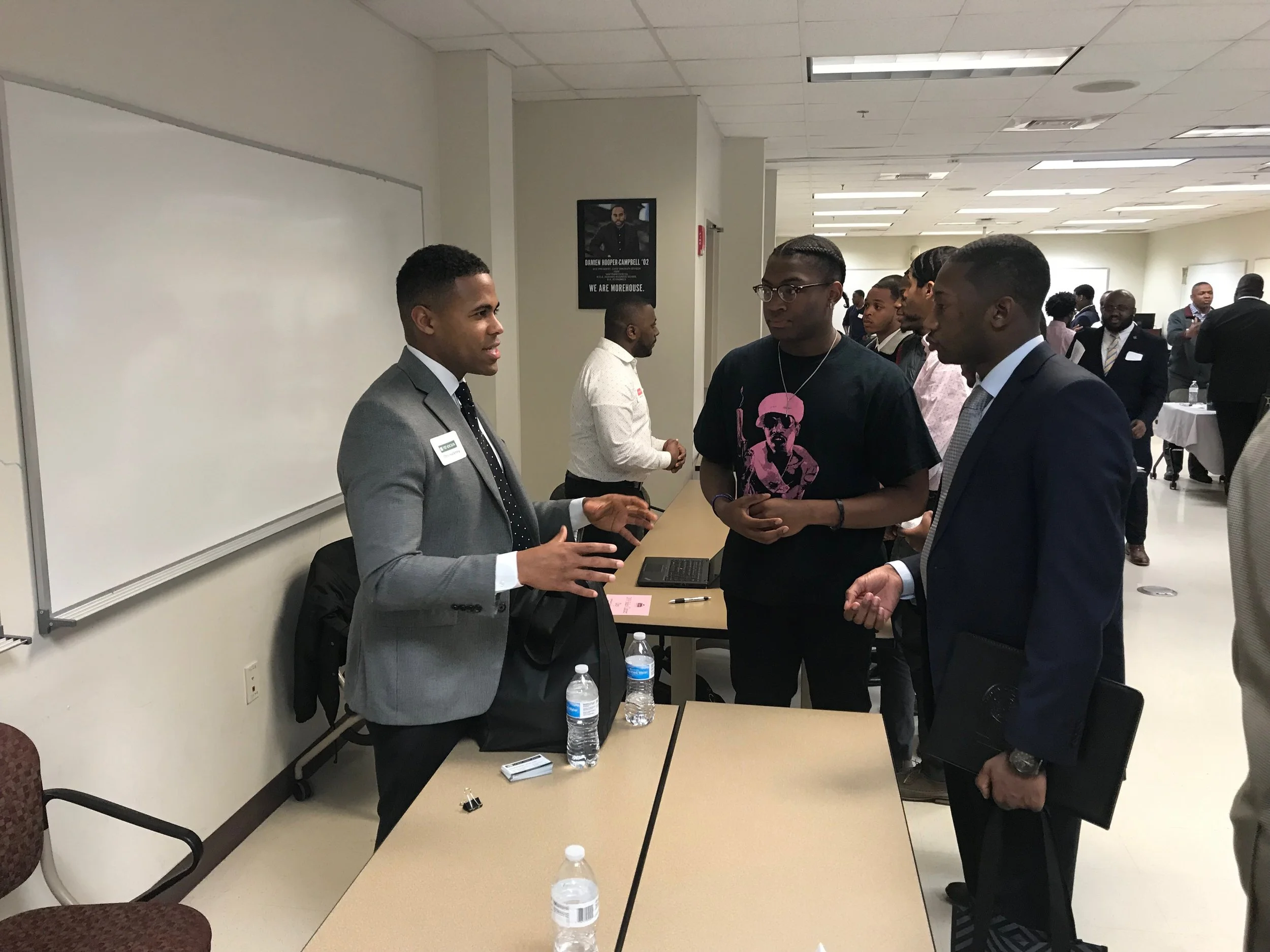 Three men in formal and casual attire engaging in conversation at a table with water bottles in a professional setting, with more people in the background.